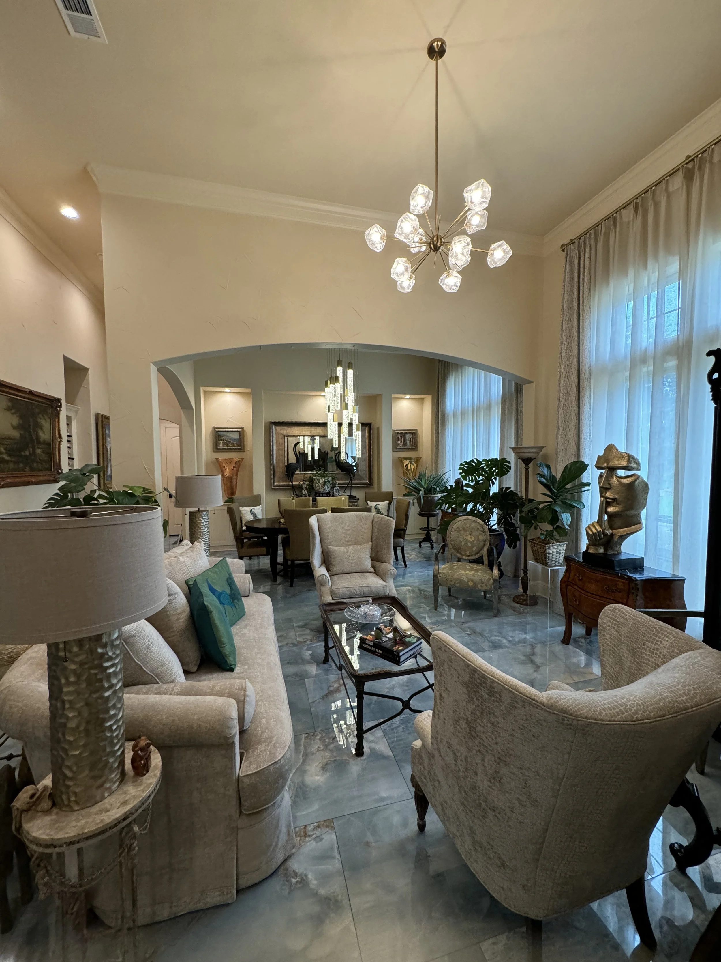 A spacious living room with beige upholstered armchairs and sofas, a glass coffee table, various decorative plants, artworks on the walls, and a large window with sheer curtains. A modern chandelier hangs from the ceiling.