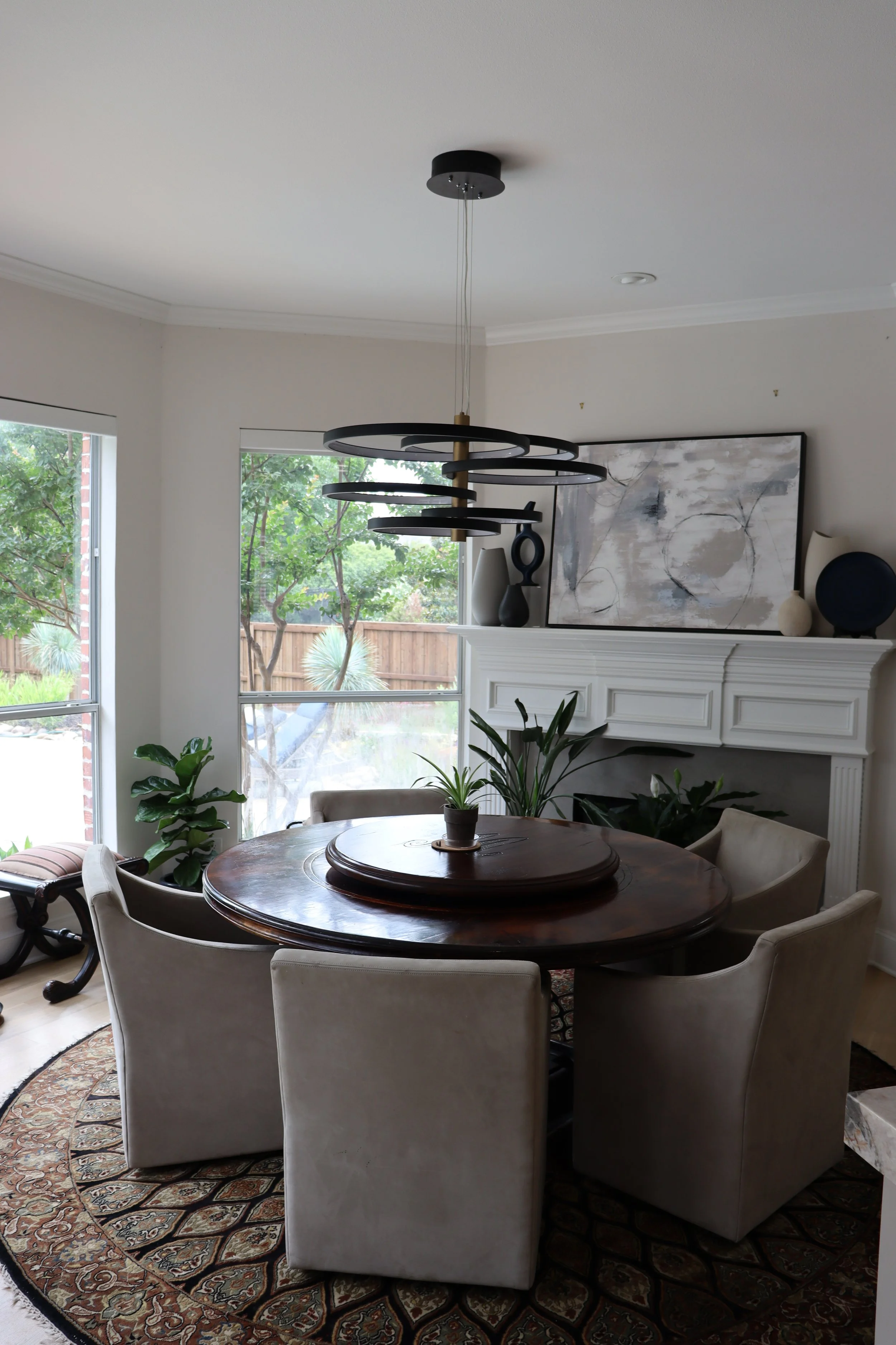 A cozy dining area with a round wooden table surrounded by six beige upholstered chairs, an ornate patterned area rug, large windows letting in natural light, green plants, a modern chandelier hanging from the ceiling, and a white mantel with abstract artwork and vases on top.