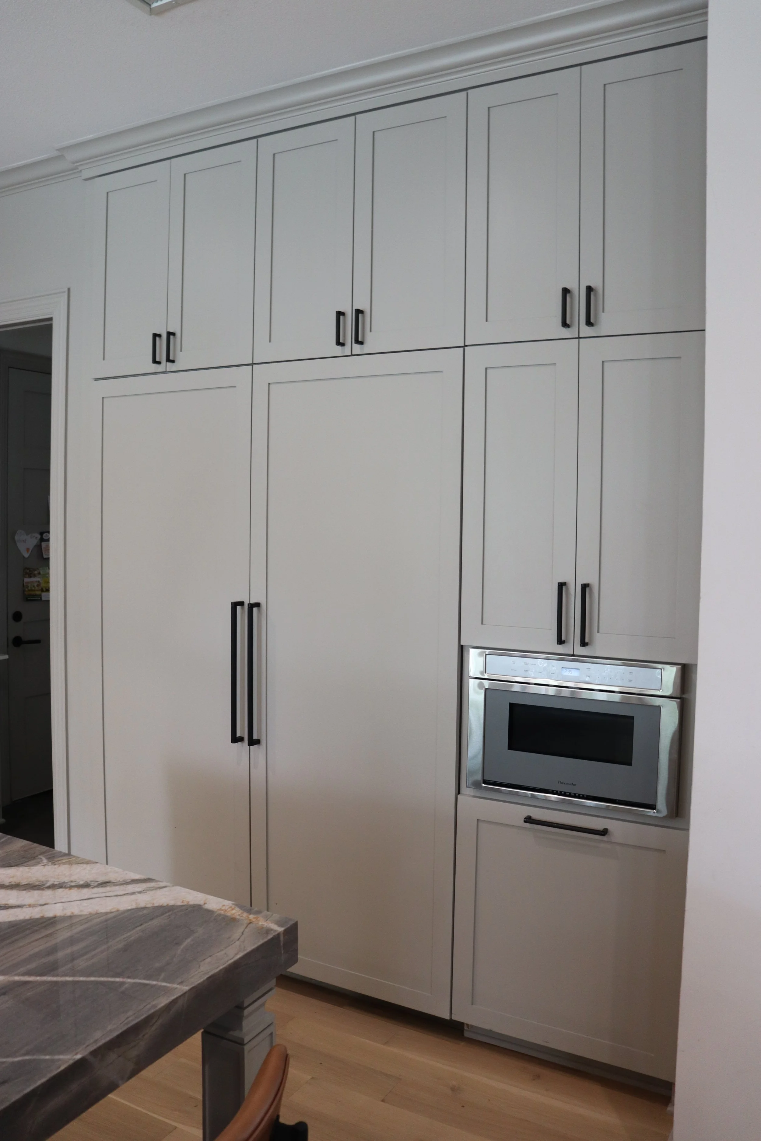 White kitchen cabinets with black handles, built-in stainless steel microwave oven, and a partial view of a wooden table with a chair.