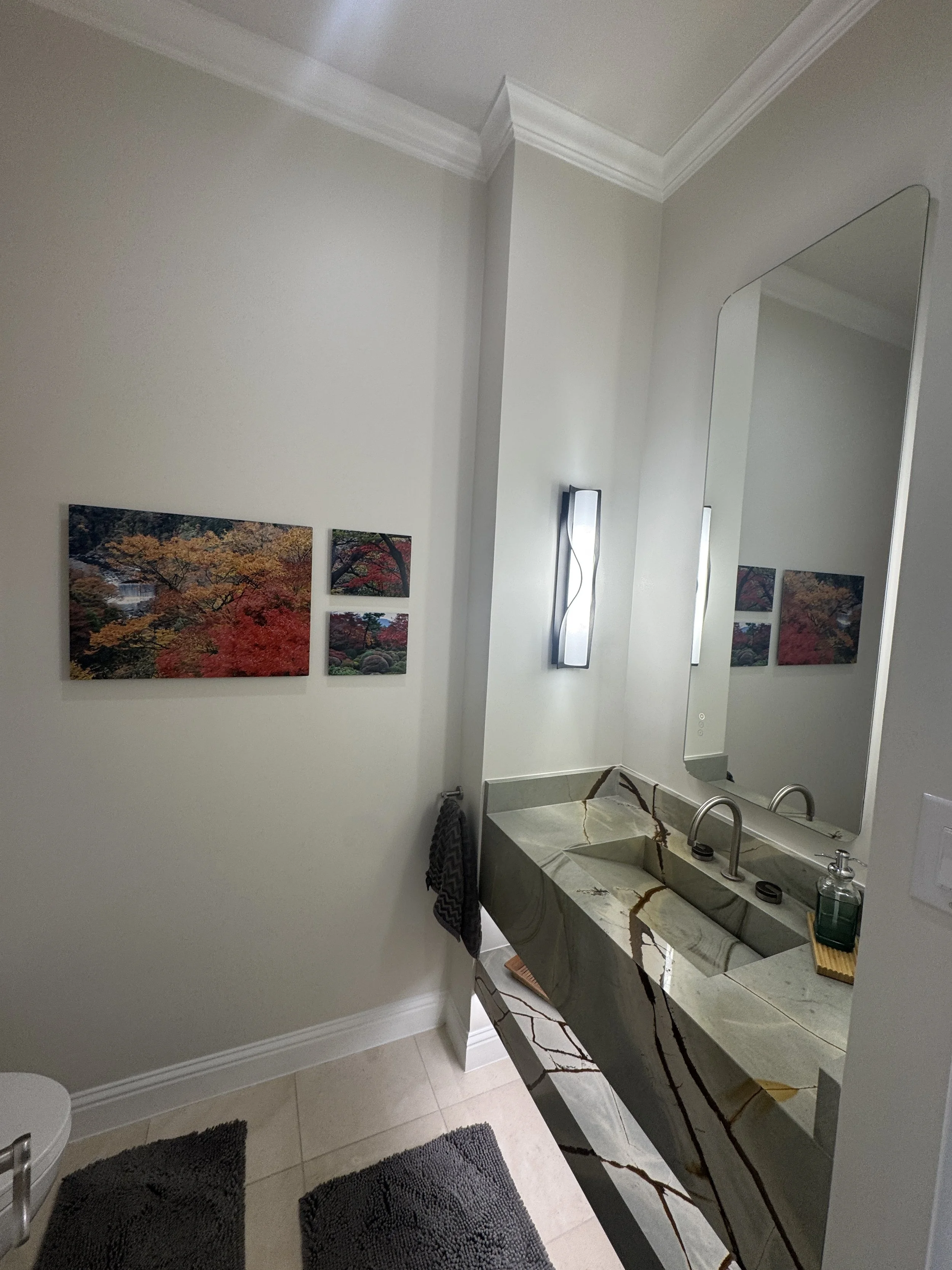 Bathroom with a modern marble sink, a large mirror, wall art of fall foliage, and a dark towel hanging on a hook.
