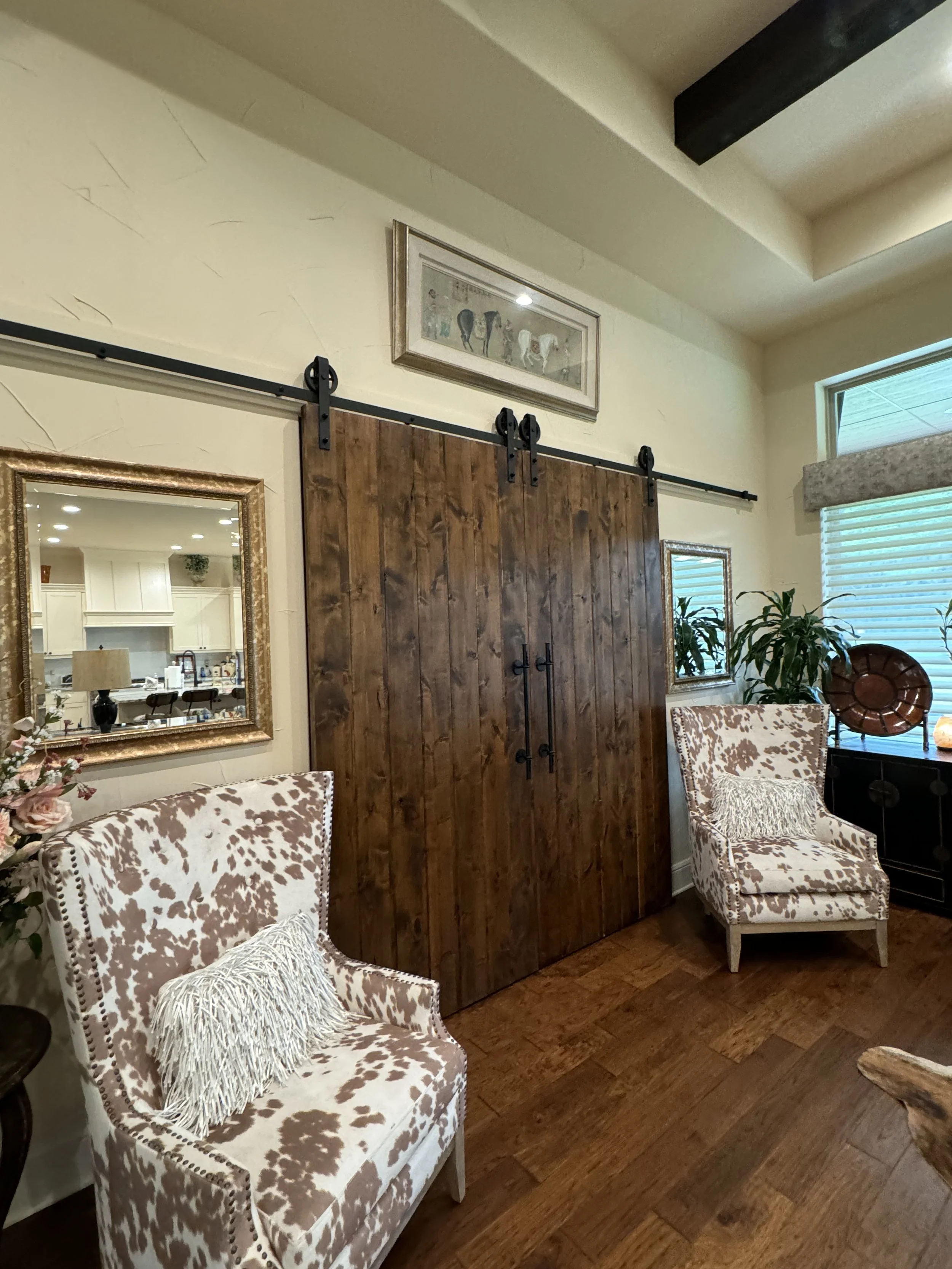 Living room with two patterned armchairs, a wooden sliding barn door, a mirror with a gold frame, plants, and a framed picture of horses on the wall.