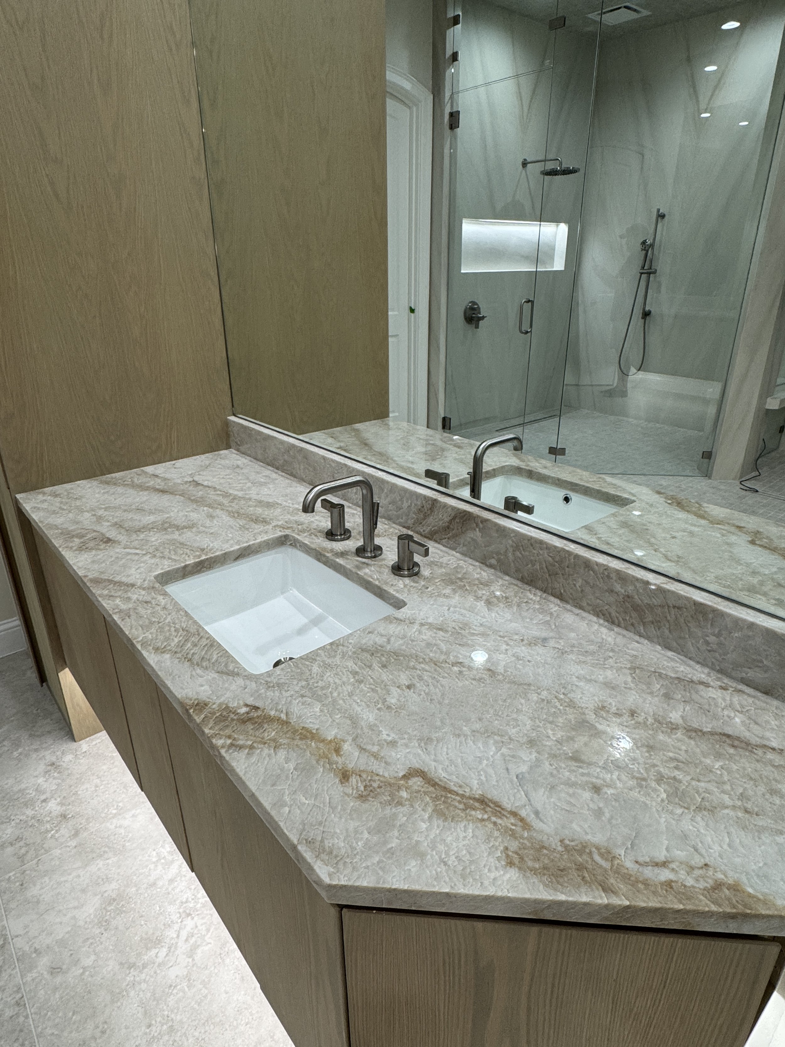 A bathroom vanity with a marble countertop, a built-in rectangular sink, and a large mirror reflecting a glass-enclosed shower area with a showerhead, a handheld shower, and a narrow window.