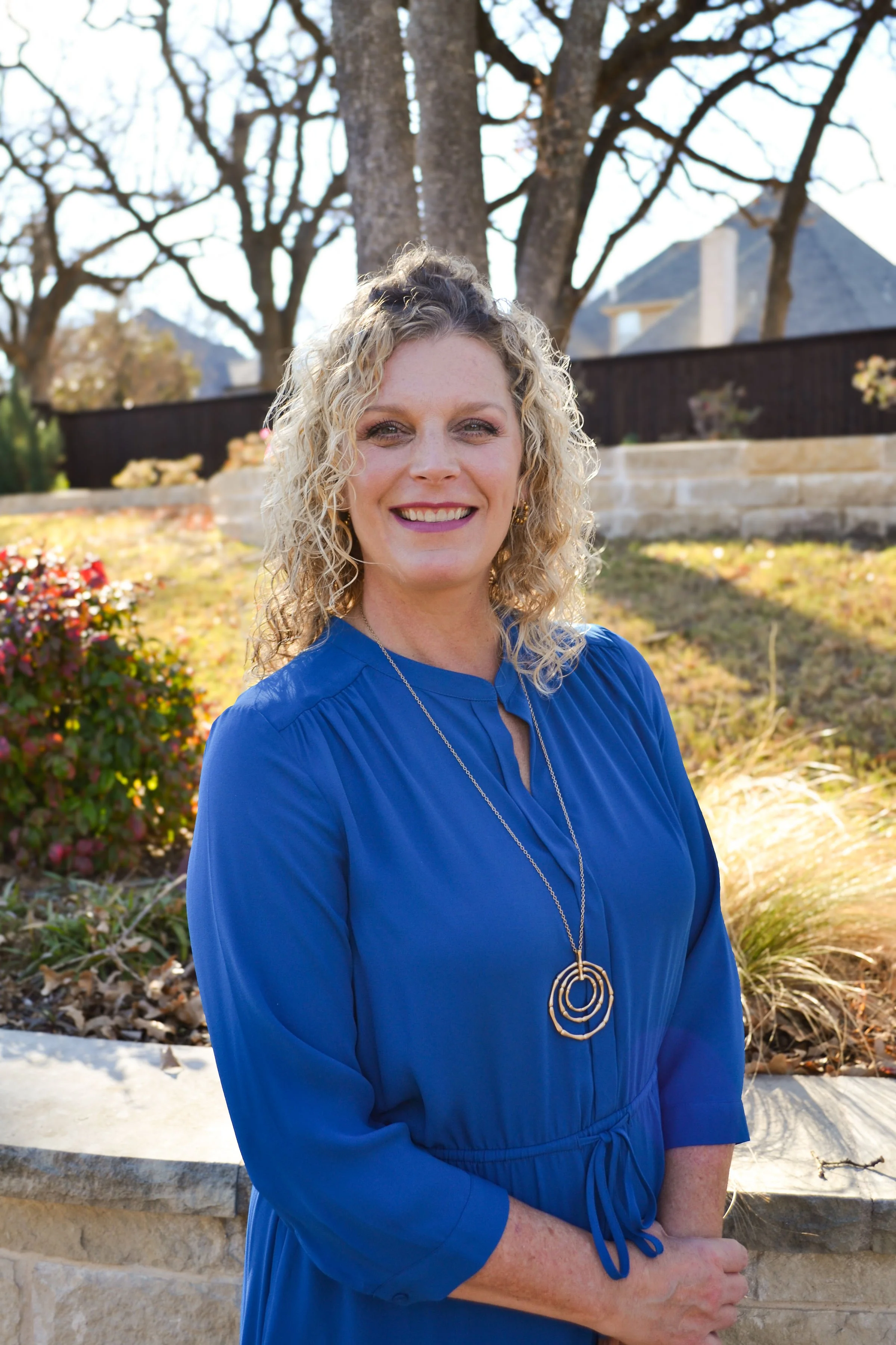 A smiling woman with curly blonde hair standing outdoors in front of trees and a stone garden wall, wearing a bright blue dress and a long necklace.