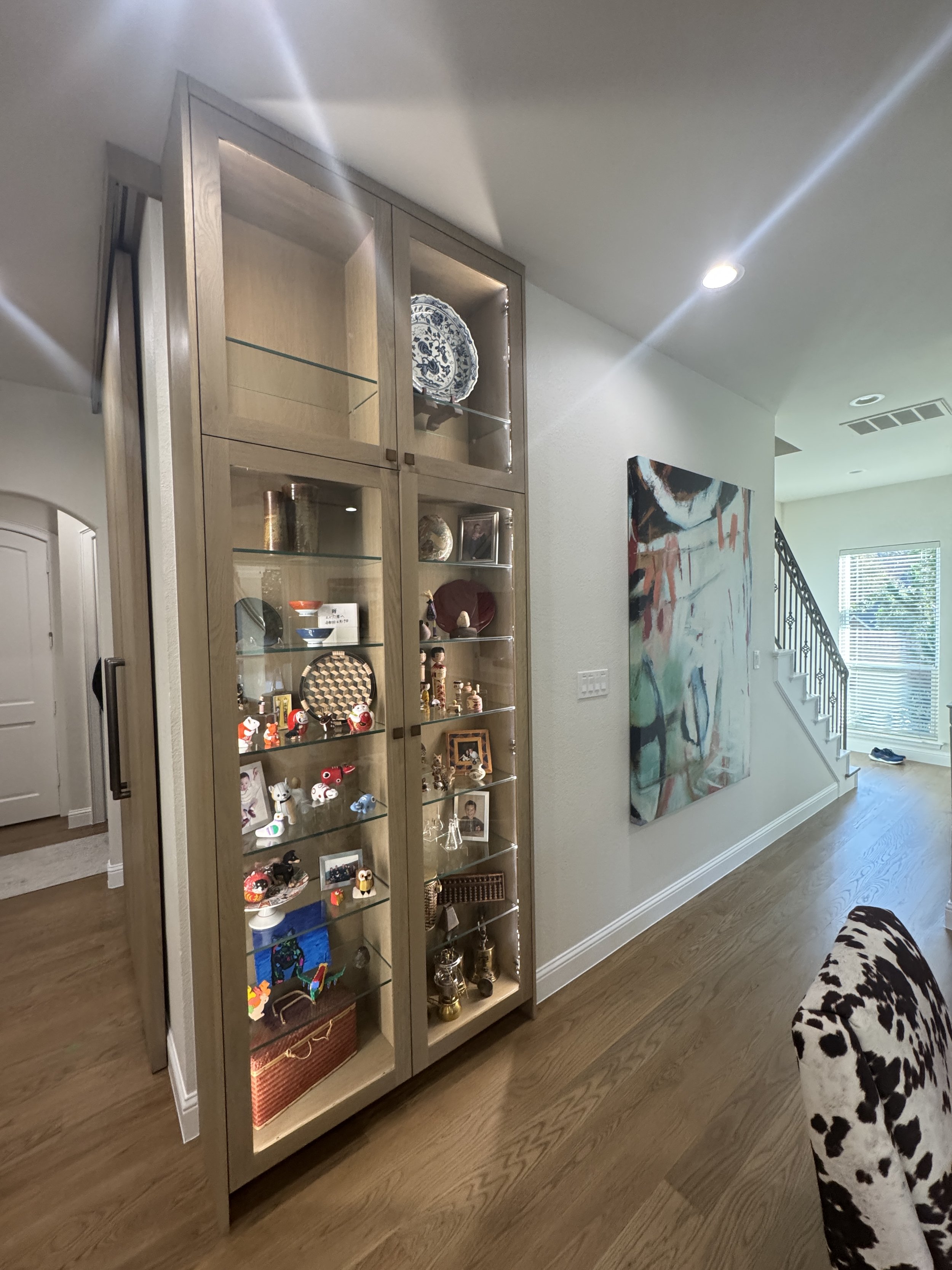 A beige display cabinet with glass doors filled with various decorative objects and souvenirs inside, positioned in a room with artwork on the wall, a staircase, and a hardwood floor.