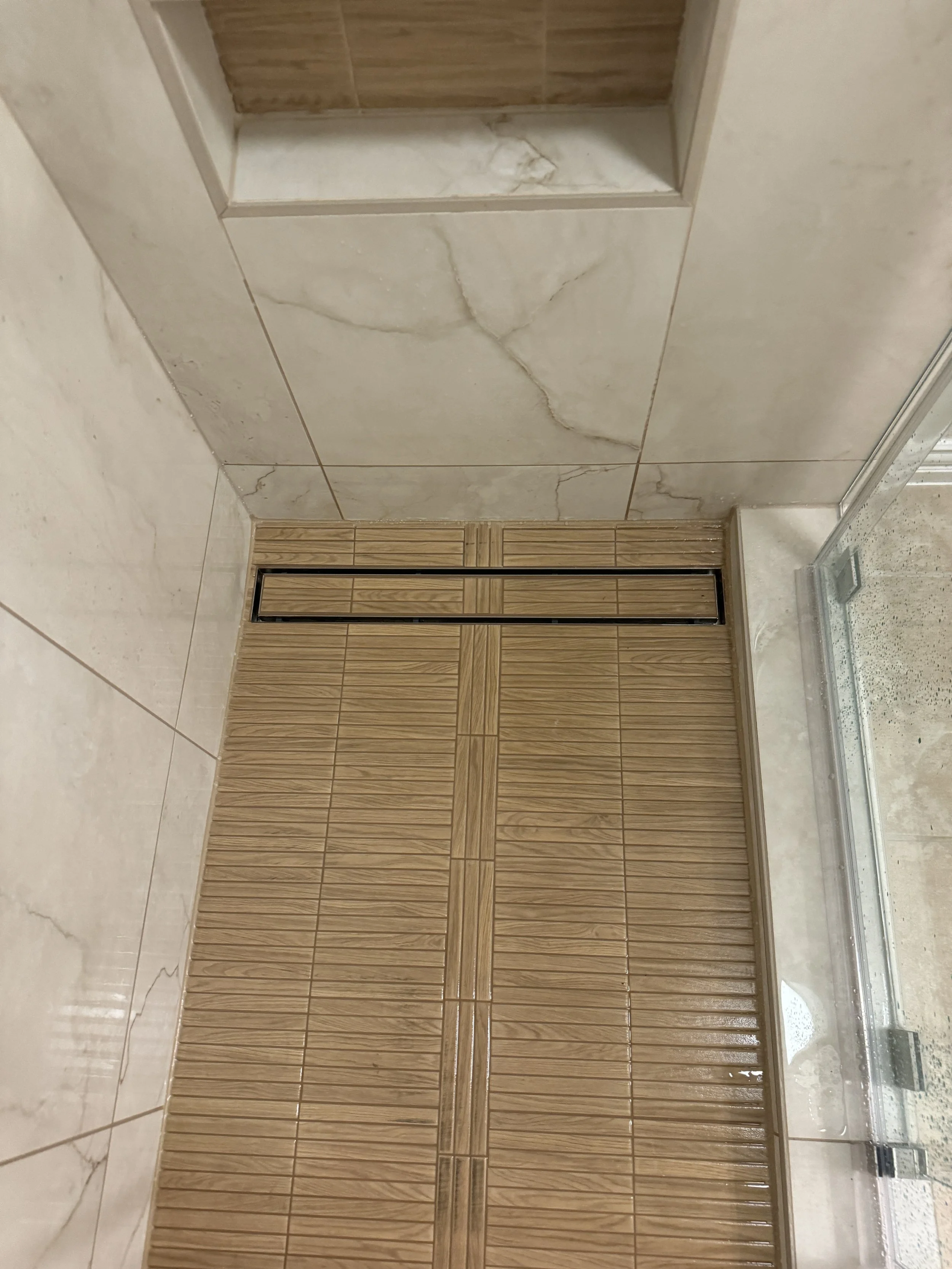 A bathroom wall with beige and brown tiles, including a section of marble tiles, a wooden door, and part of a shower area.