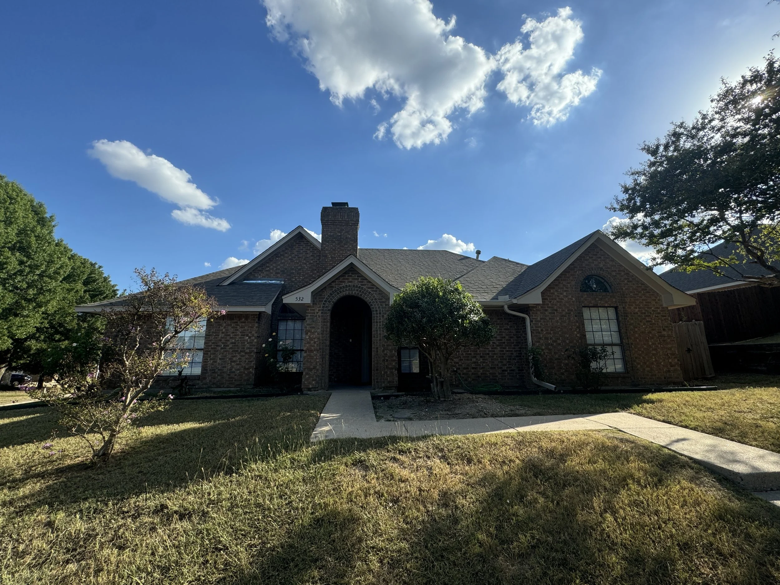 A brick house with a dark roof, a chimney, front yard with grass, a sidewalk, green trees, and a partly cloudy blue sky.
