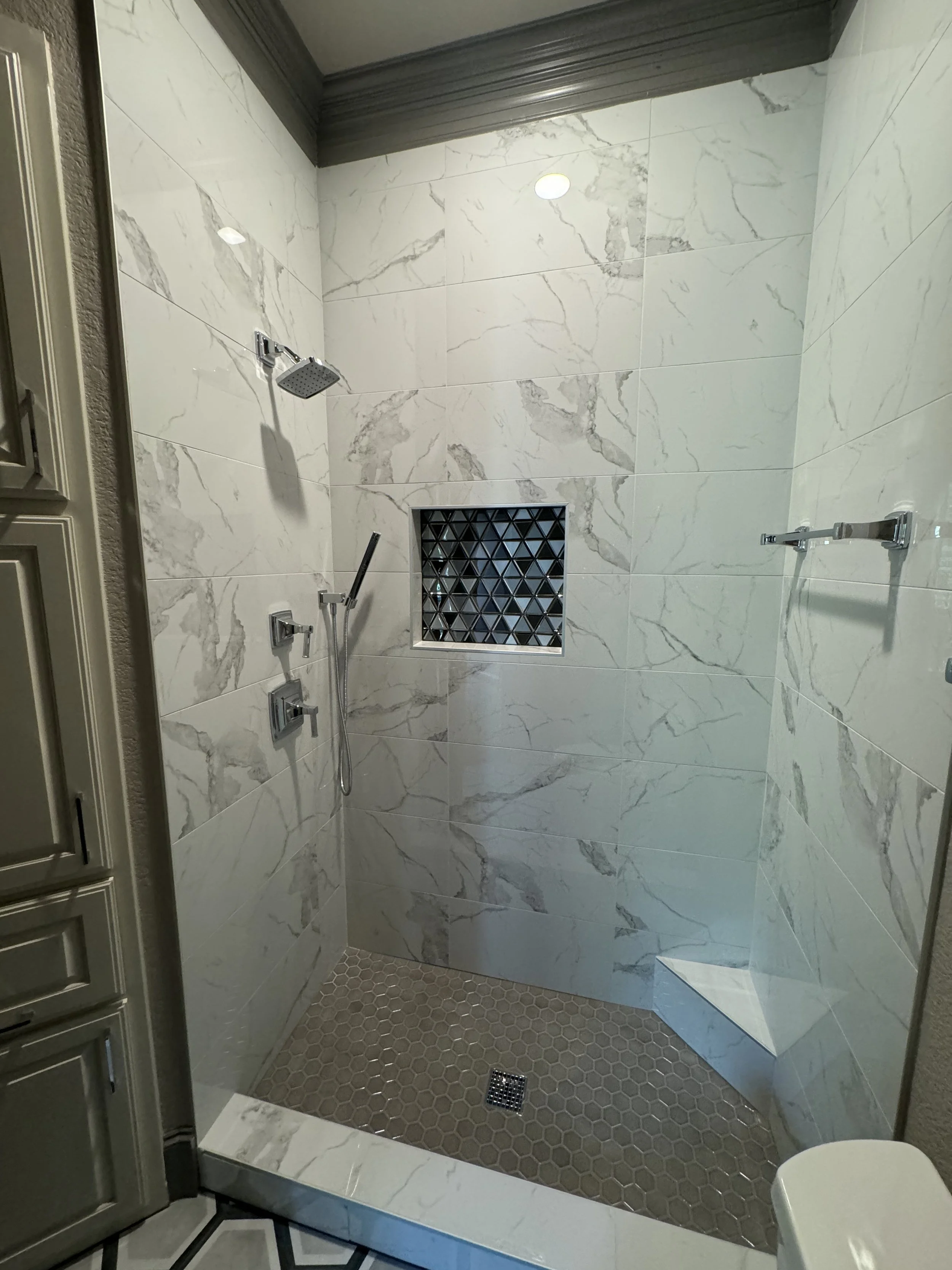 Modern walk-in shower with marble walls, a square rainfall showerhead, a hand-held shower, a small black triangular tile niche, and gray hexagonal floor tiles.