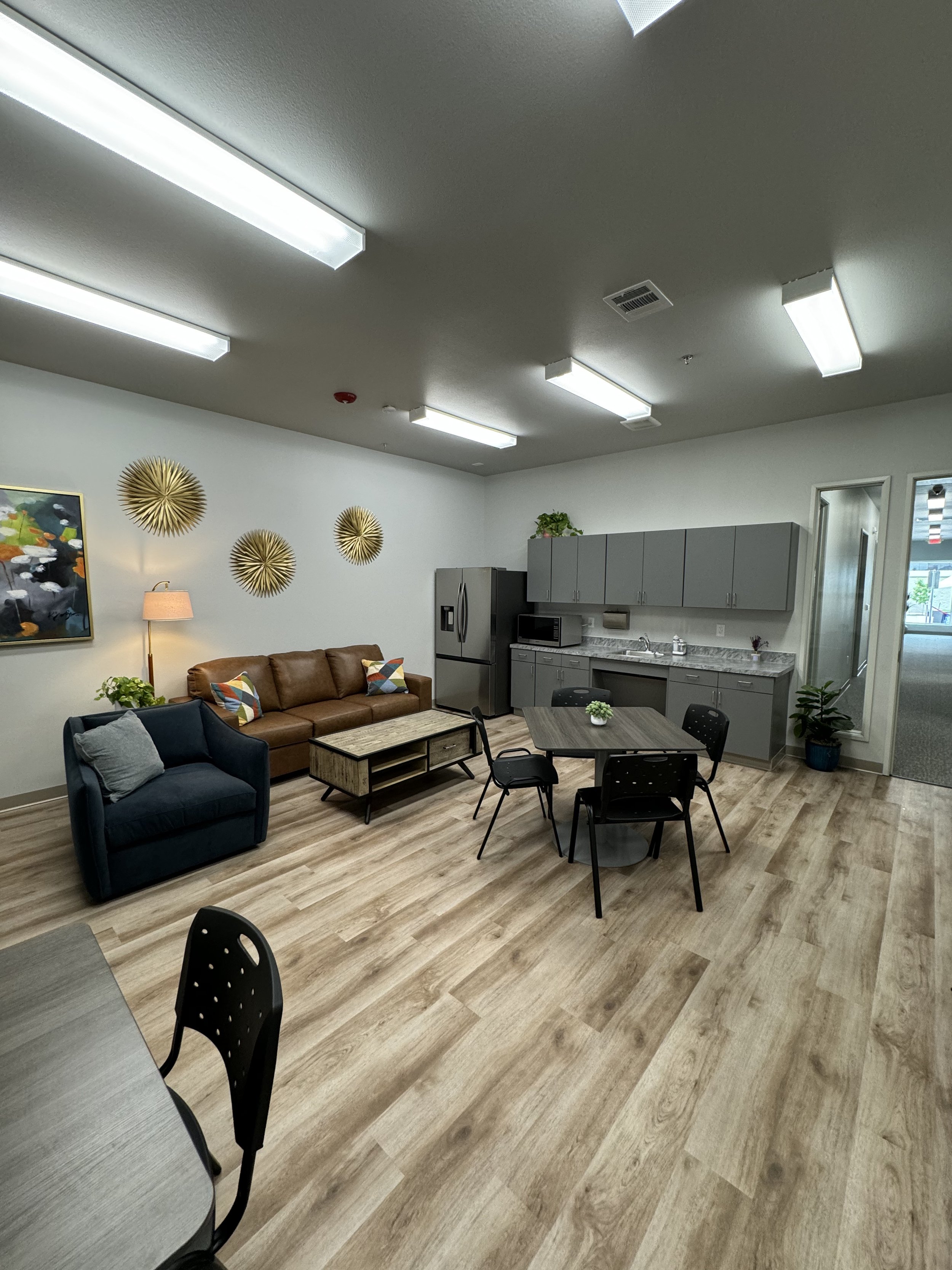 A common area in an office or break room featuring a seating arrangement with a brown leather sofa and a small black armchair, a wooden coffee table, and black chairs around a circular table. The space has wall decorations, including three gold sunburst wall art pieces, a painting, a plants, and a table lamp. The room has a kitchenette with gray cabinets, a countertop, a microwave, a refrigerator, and a sink. The ceiling has multiple fluorescent light fixtures and a view of the outdoors through a window or door.