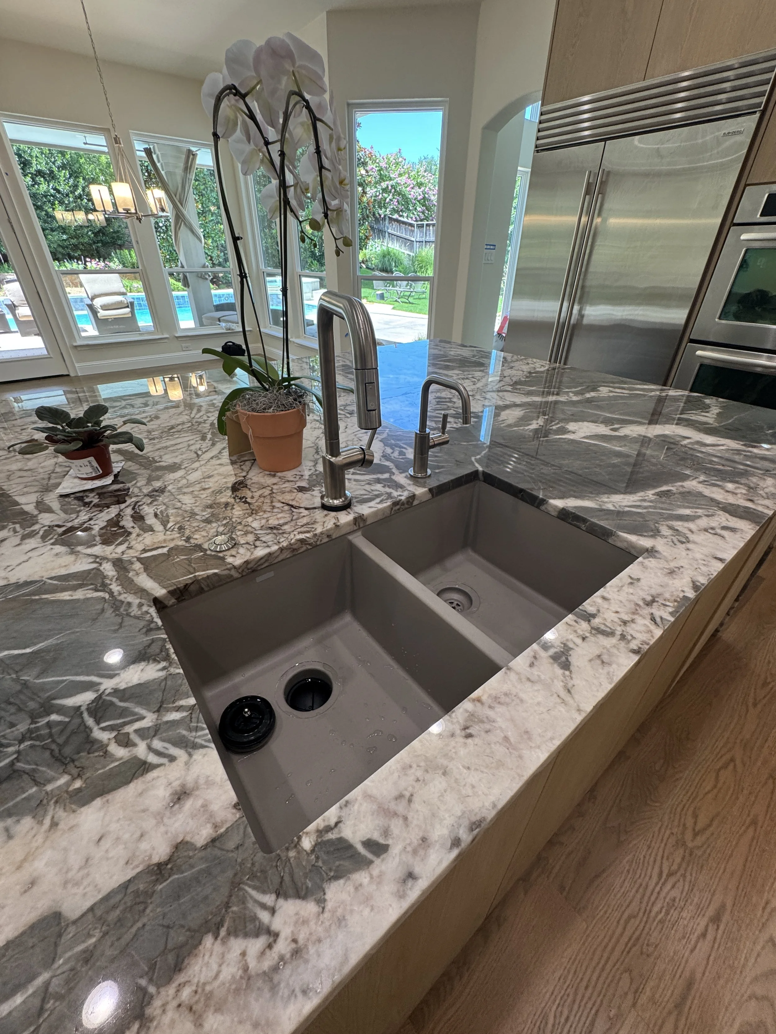 A kitchen countertop with a marble surface, a stainless steel double sink, and a modern chrome faucet. Two potted plants rest on the counter near the sink. Large windows and glass doors behind reveal an outdoor patio with seating, greenery, and a swimming pool.