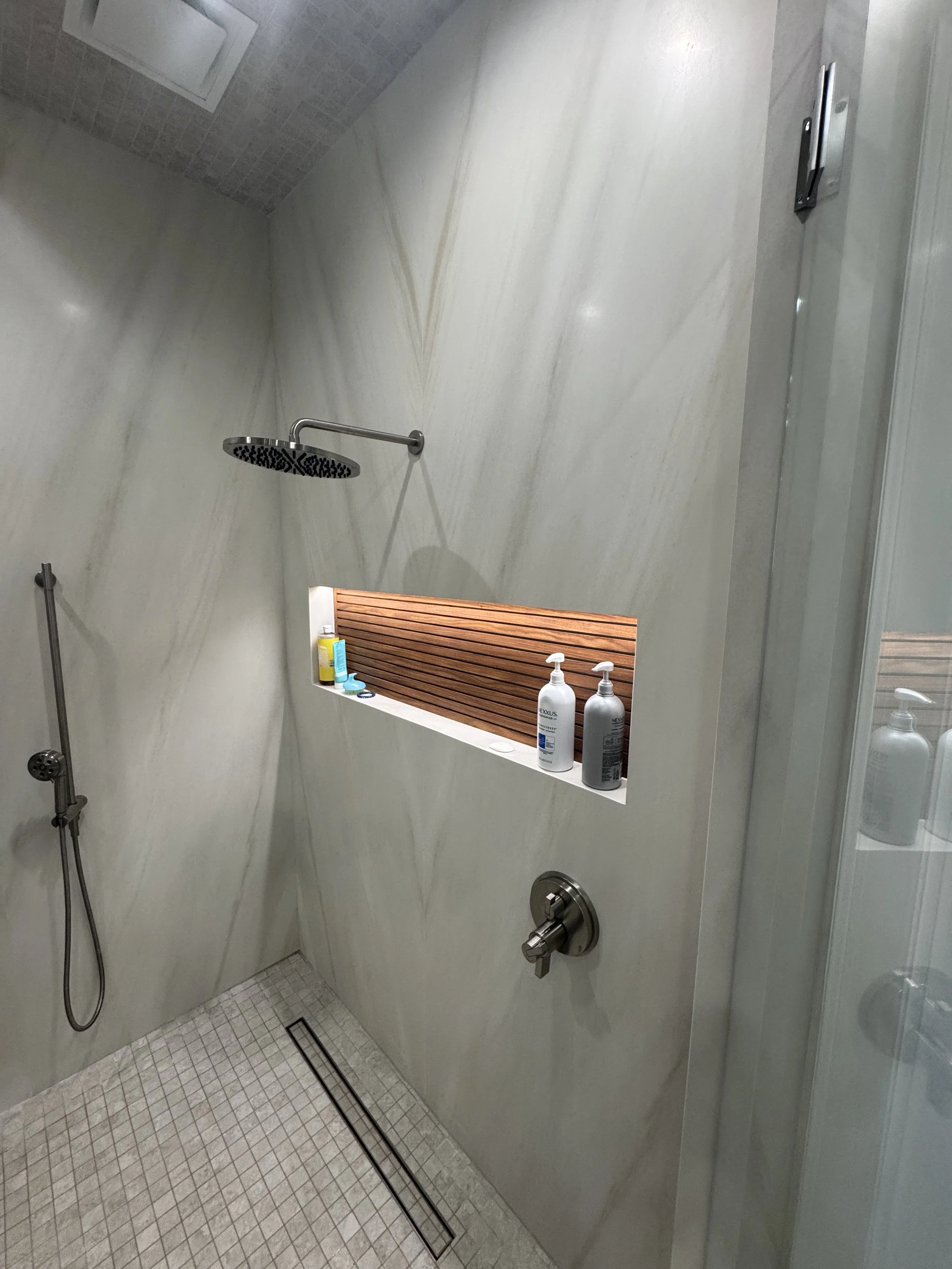 Bathroom shower area with a rainfall showerhead, handheld shower, and built-in shelf holding toiletries, with tile walls and floor, and a window on the wall.