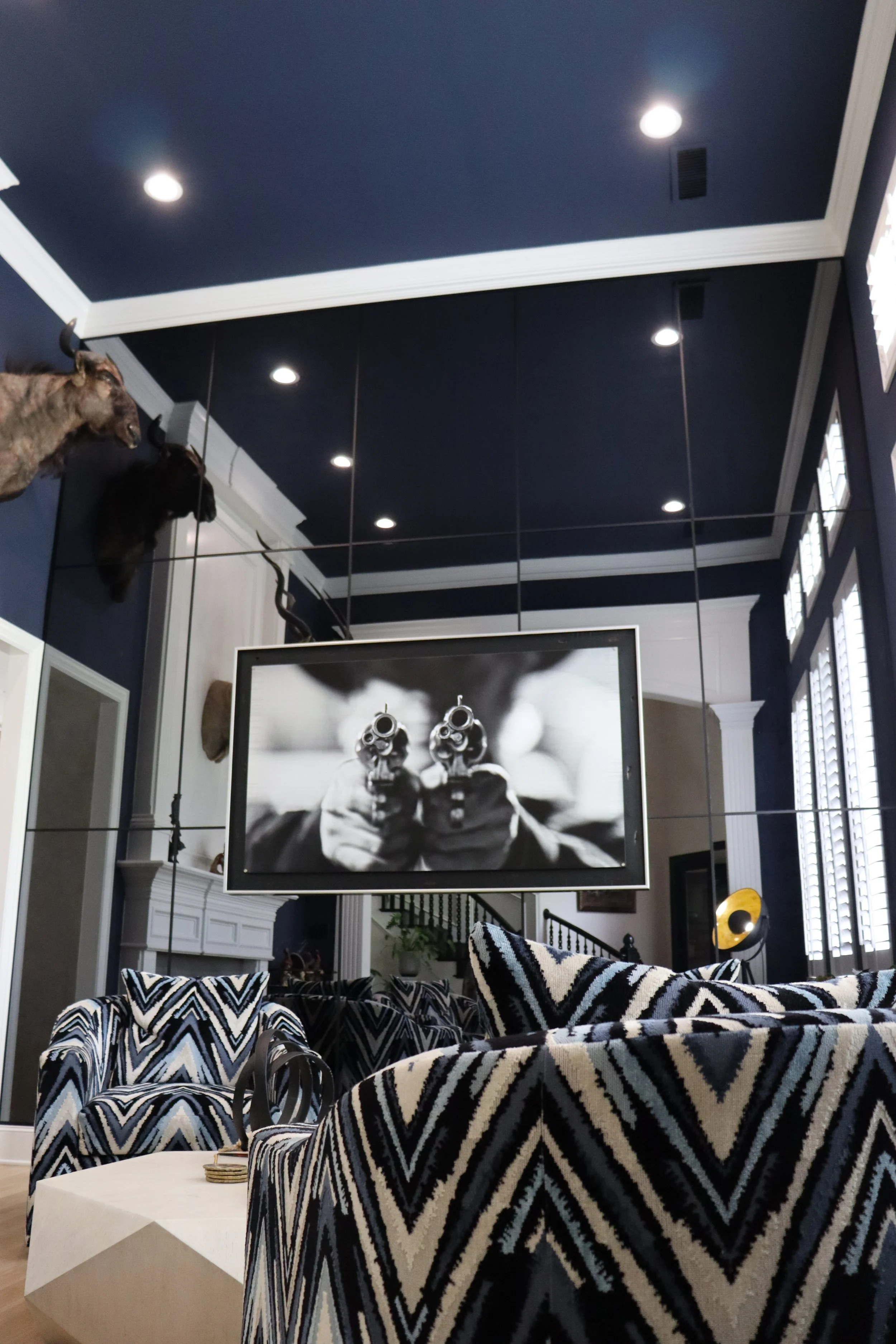 Living room with dark blue walls and ceiling, decorated with mounted animal heads, including a wolf and a goat. A large black-and-white photograph of a person aiming a gun is displayed on a screen. The room has modern furniture, including patterned armchairs, a white geometric coffee table, and floor-to-ceiling windows with blinds.
