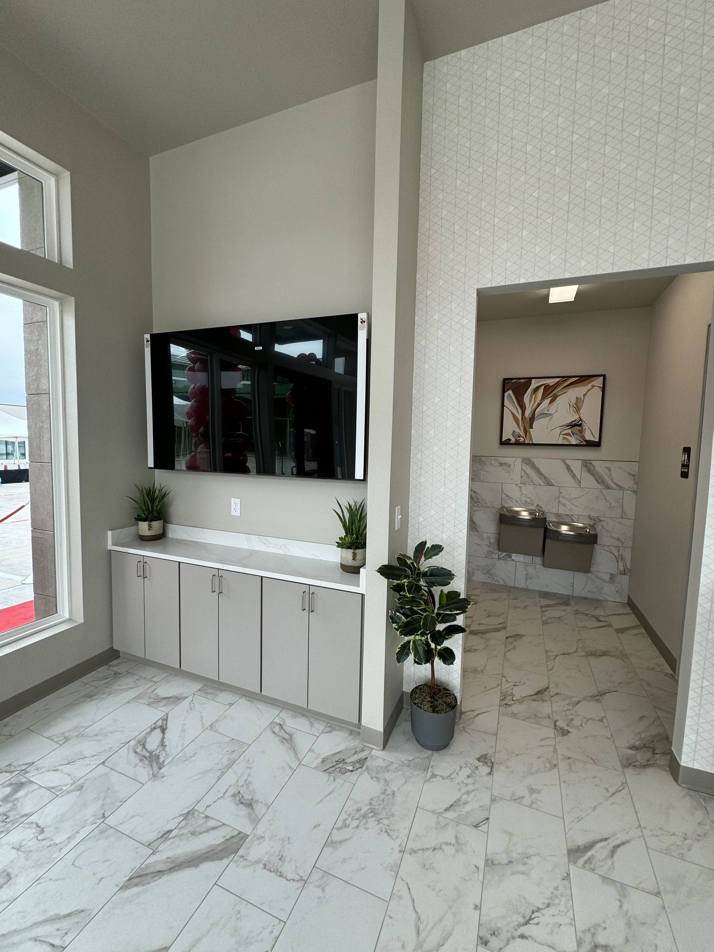 Interior of a modern building with marble flooring, a white cabinet, potted plants, a wall-mounted TV, artwork, and a hallway with two trash bins.