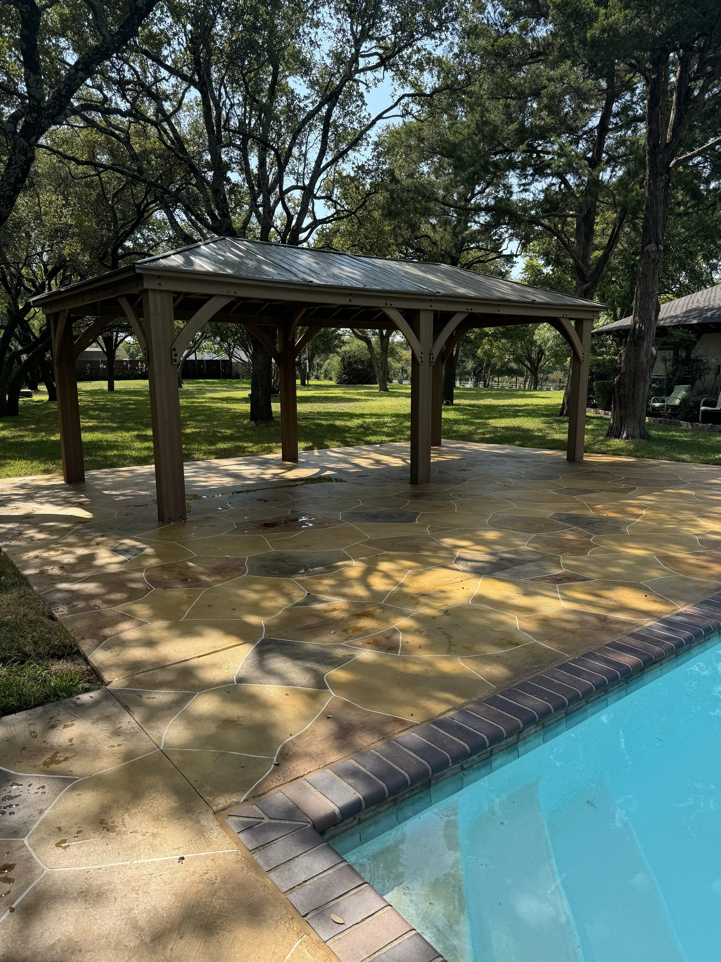 Backyard scene with a wooden gazebo, a swimming pool with water, green grass, and trees providing shade.