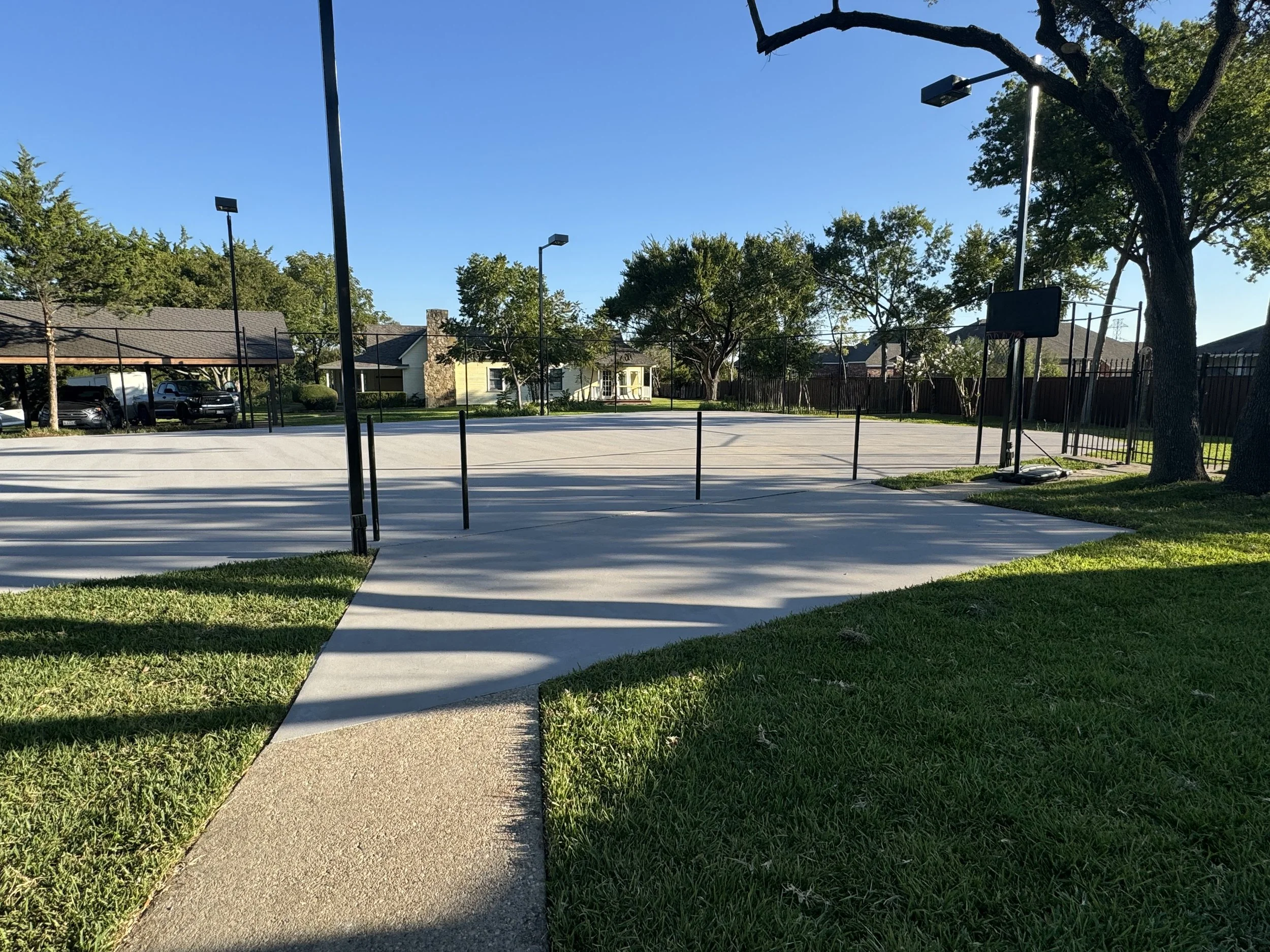 An outdoor basketball court surrounded by a black metal fence, with four tall light poles, trees, and houses in the background. The court has a hoop on a stand, and the area is illuminated by natural sunlight with shadows cast on the ground.