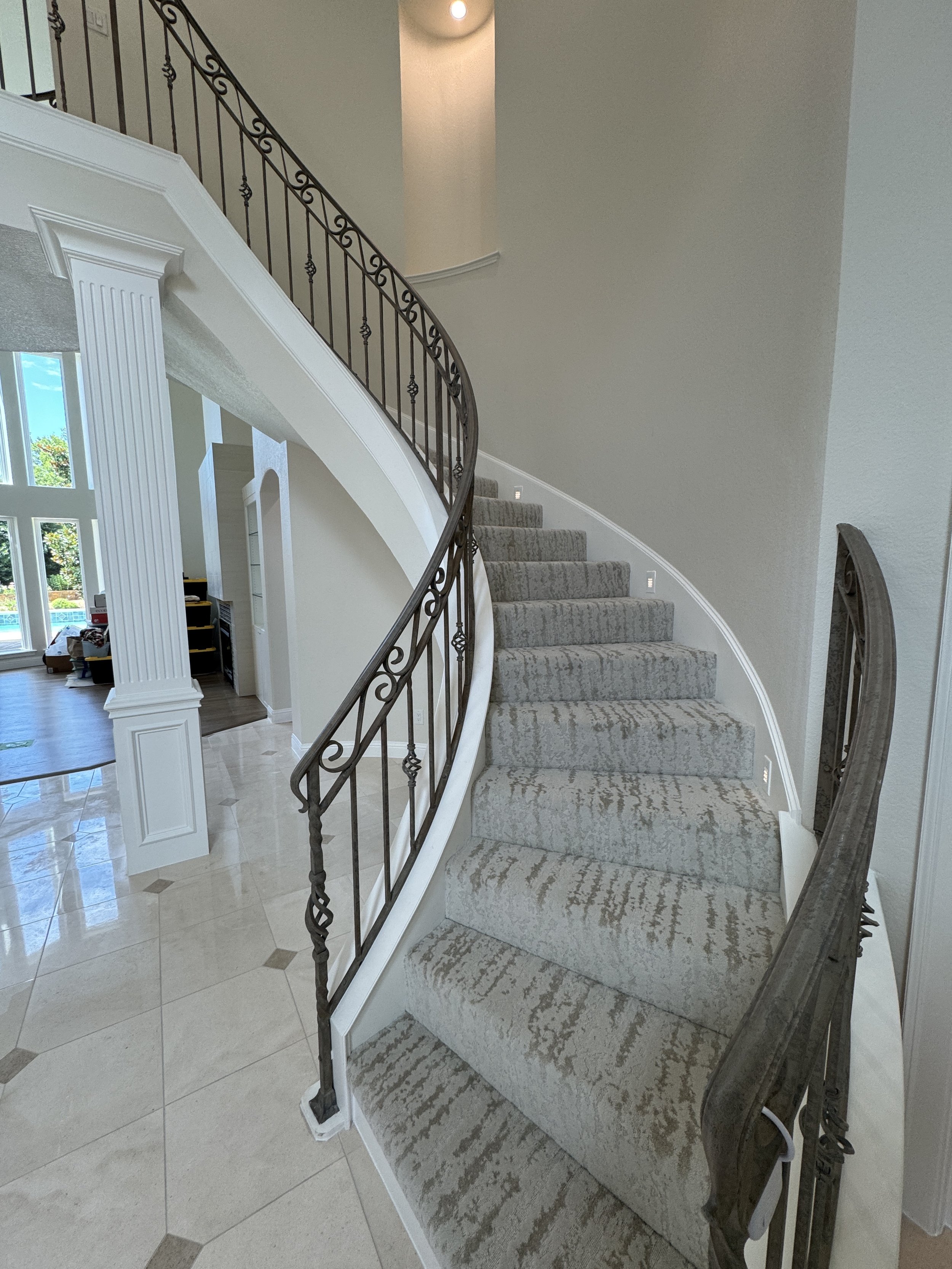Interior view of a curved staircase with a wrought iron railing, beige carpeted stairs, and a light-colored wall with ambient lighting.
