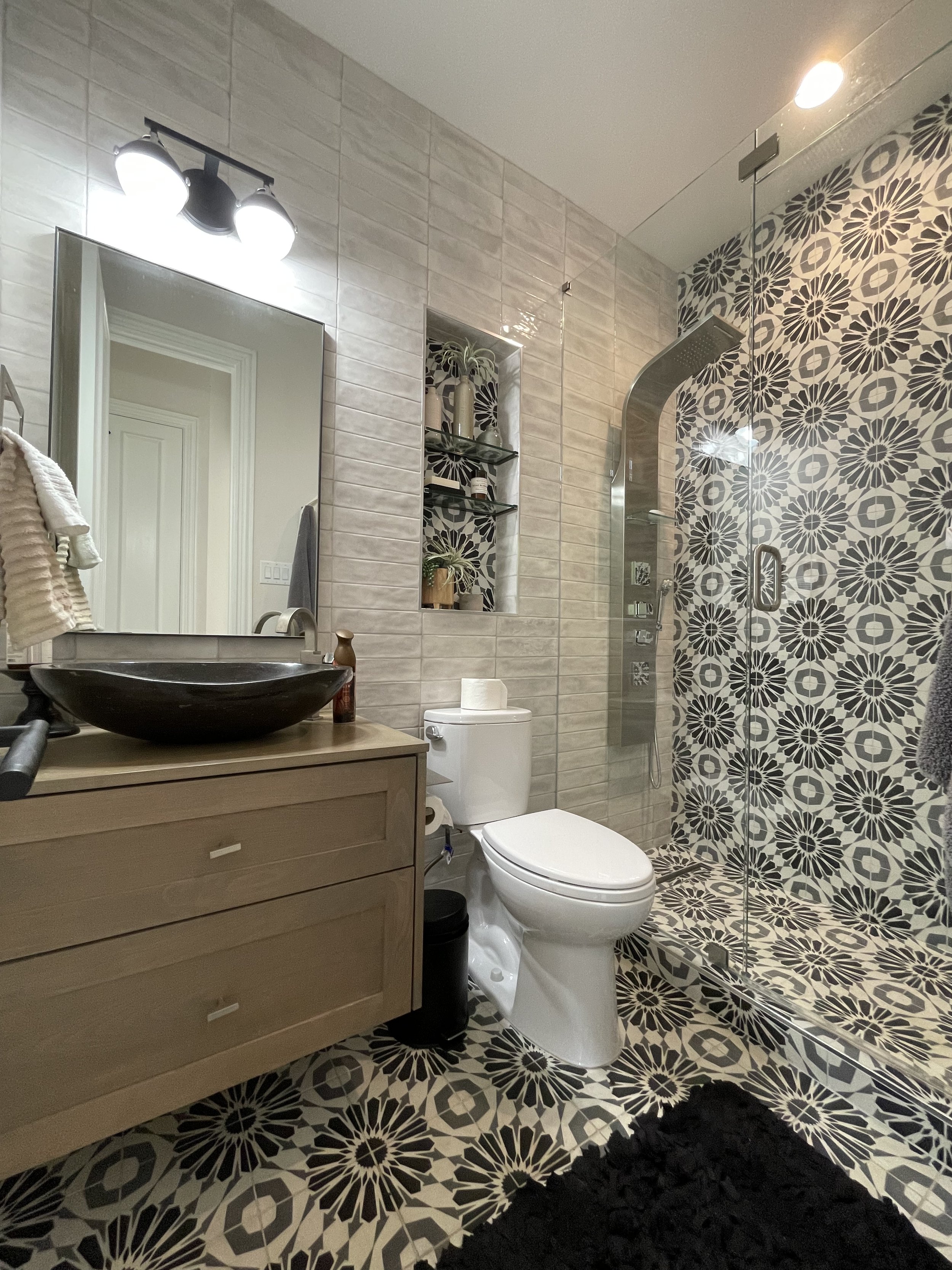Modern bathroom with a patterned tile shower wall, a white toilet, a wooden vanity with a black vessel sink, a mirror, and wall shelves with decorative items.