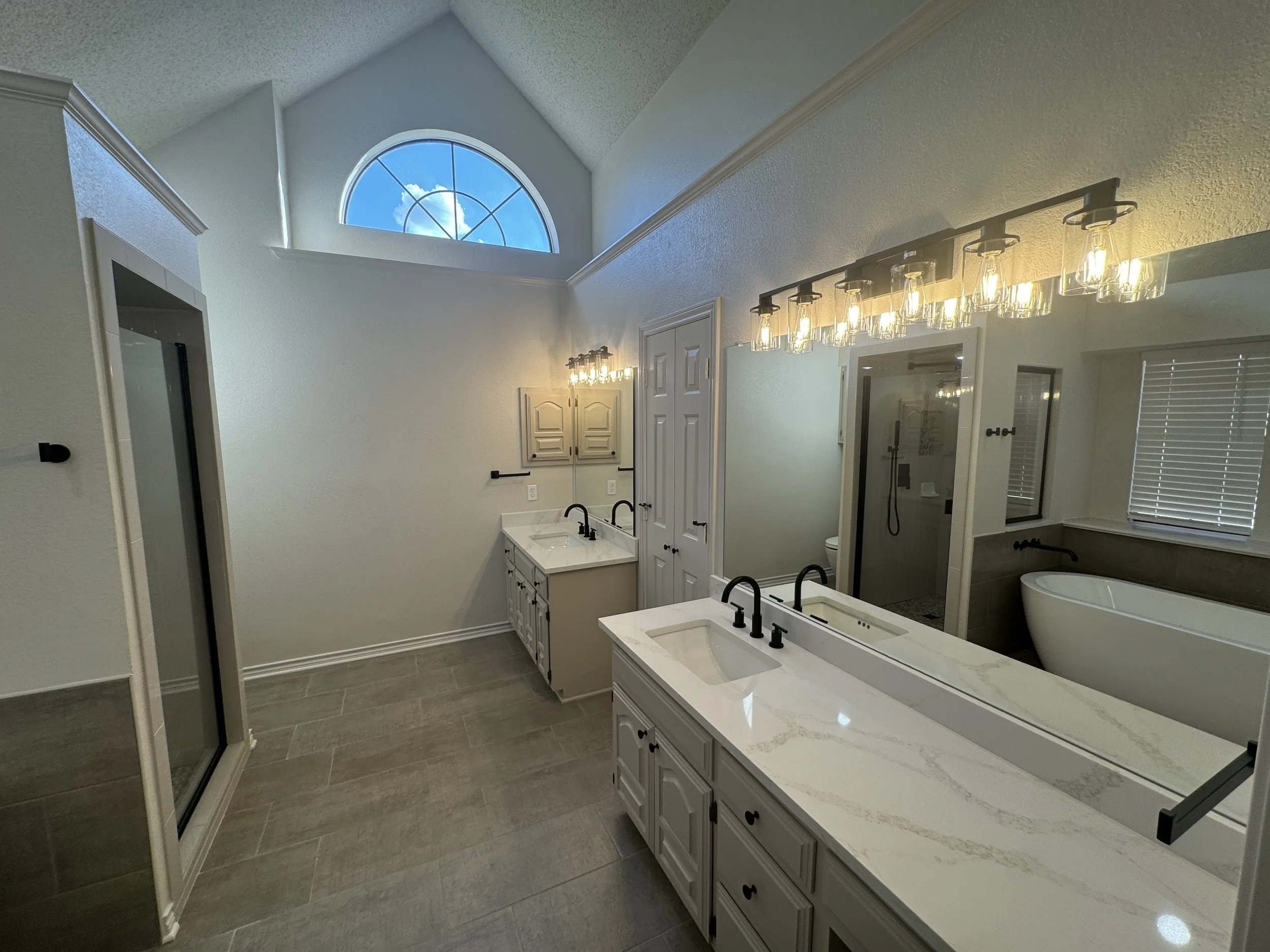 Interior view of a modern bathroom with white cabinets, double sink, large mirror, wall-mounted lighting, and a bathtub, with a large arched window showing a blue sky and clouds.