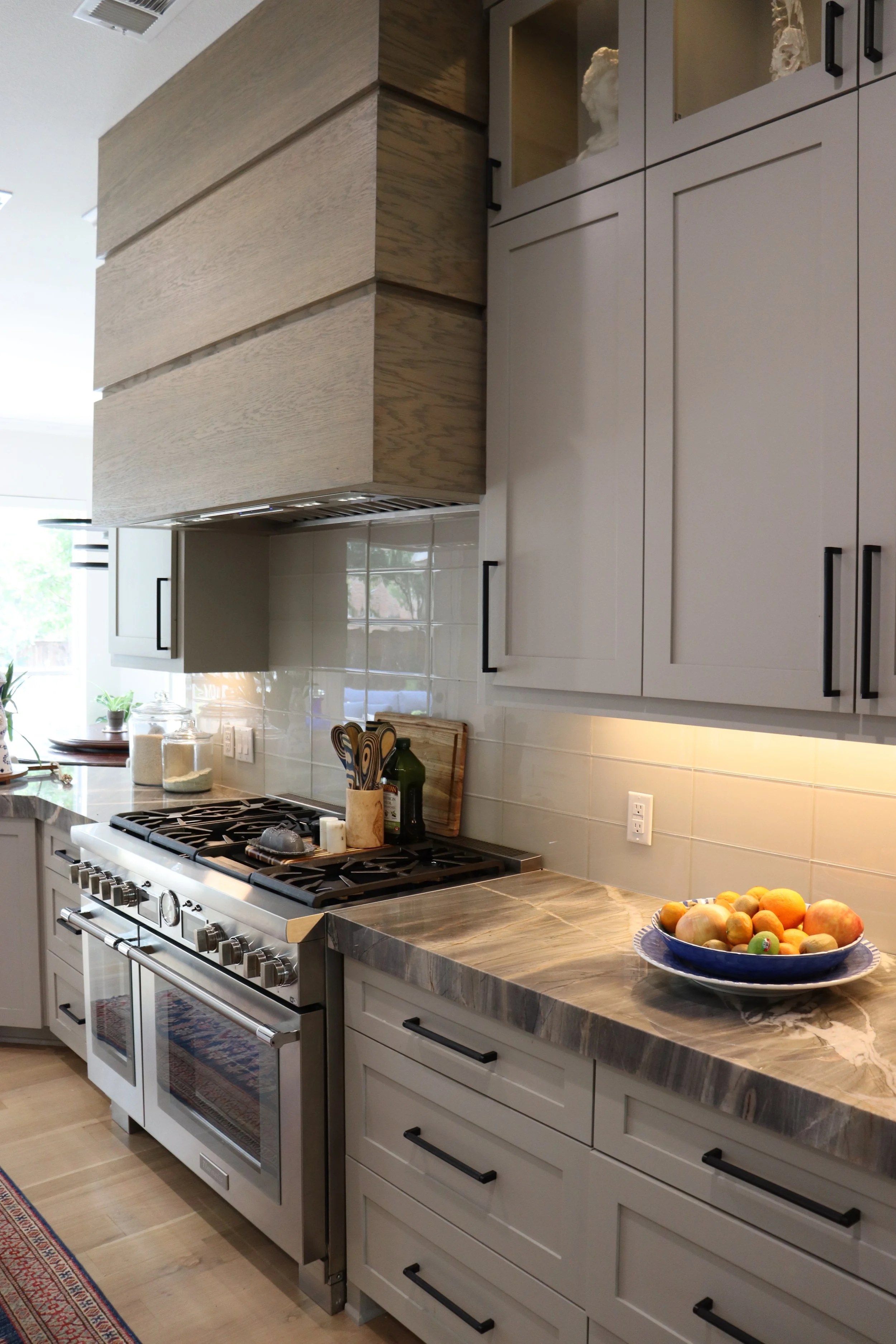 A modern kitchen with white cabinetry, a marble countertop, and a stainless steel oven and stove. There is a bowl of mixed fruits on the counter and various kitchen utensils and jars nearby. The space is well-lit with natural light.