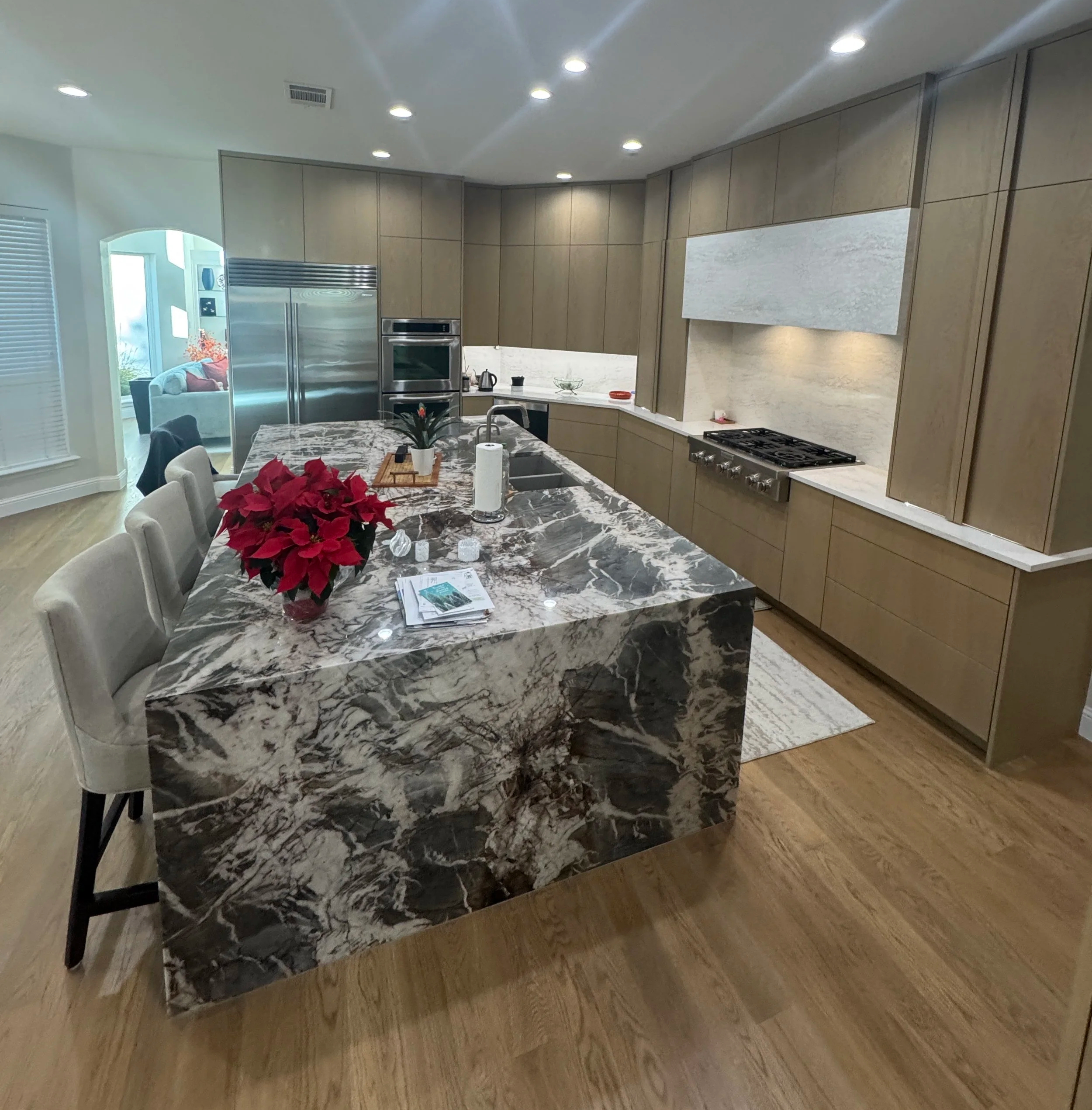 A kitchen with beige cabinets, a marble island with a poinsettia plant, paper towels, and a paper on the counter, stainless steel appliances, and a dining area with a white couch and a window with blinds.