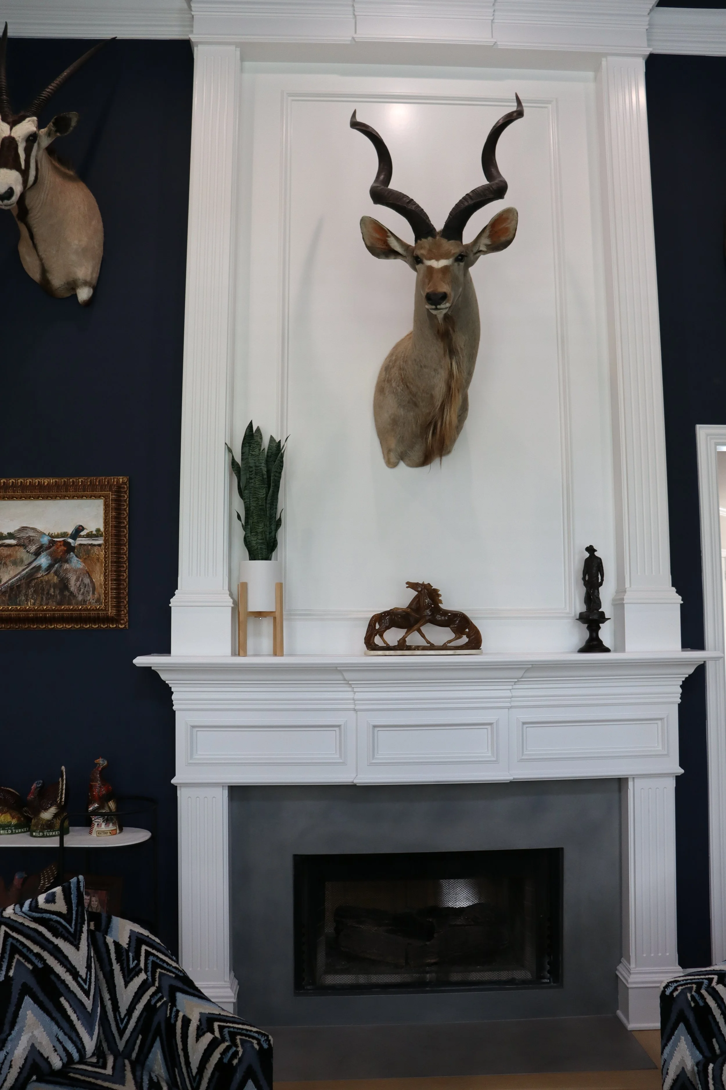 A living room with a white fireplace mantel and a mounted deer head with large antlers on a white panel above the fireplace. To the left of the fireplace is a navy blue wall with a mounted gazelle head. Below is a white shelf with decorative items, including small horse sculptures and a framed picture of a bird. In front of the fireplace are two armchairs with black, white, beige, and blue geometric patterns.