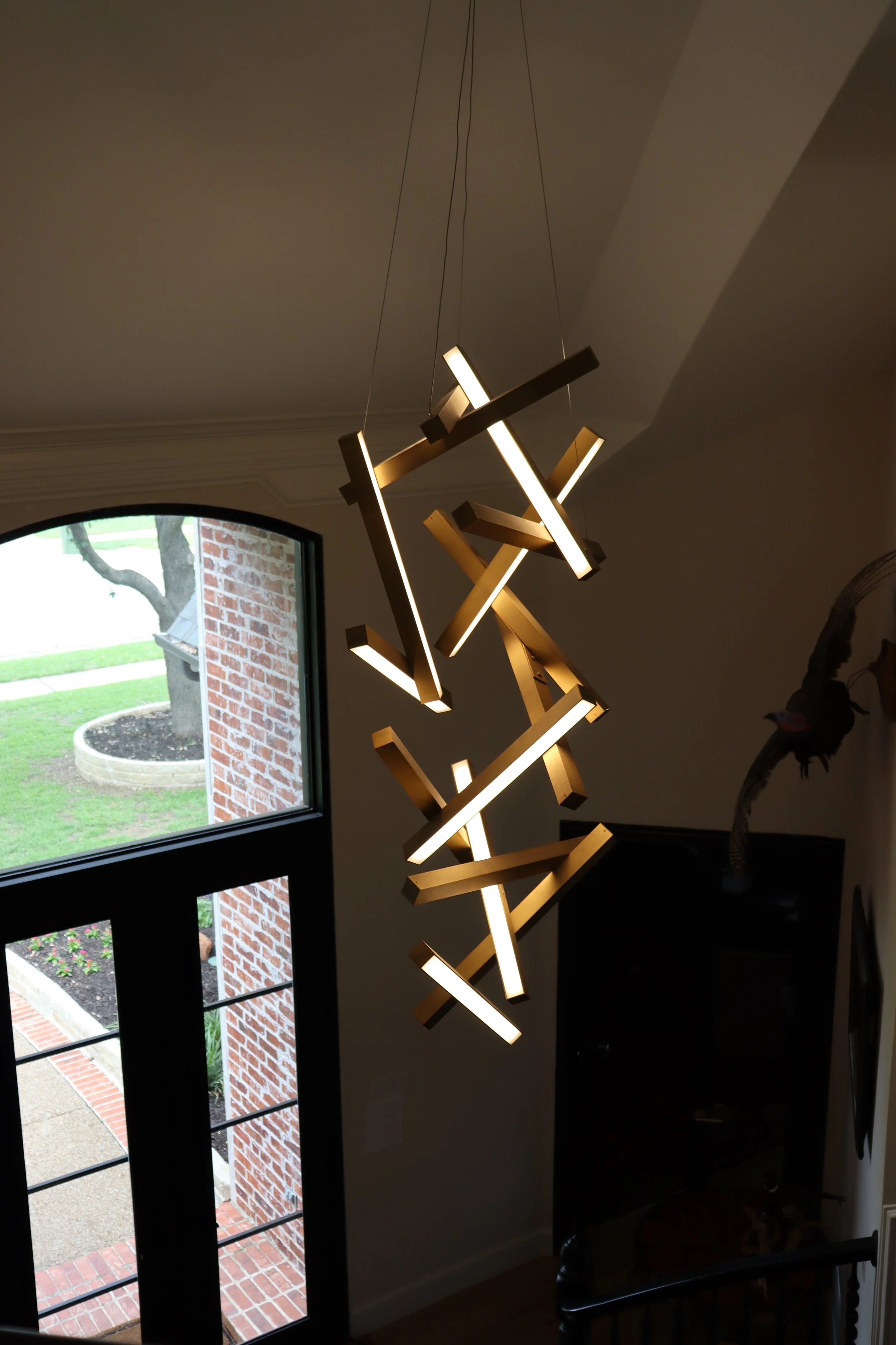 Modern hanging light fixture with multiple illuminated rods arranged in an abstract pattern, located inside a room near a window with a view of a brick-paved walkway, a tree, and a flower bed outside.
