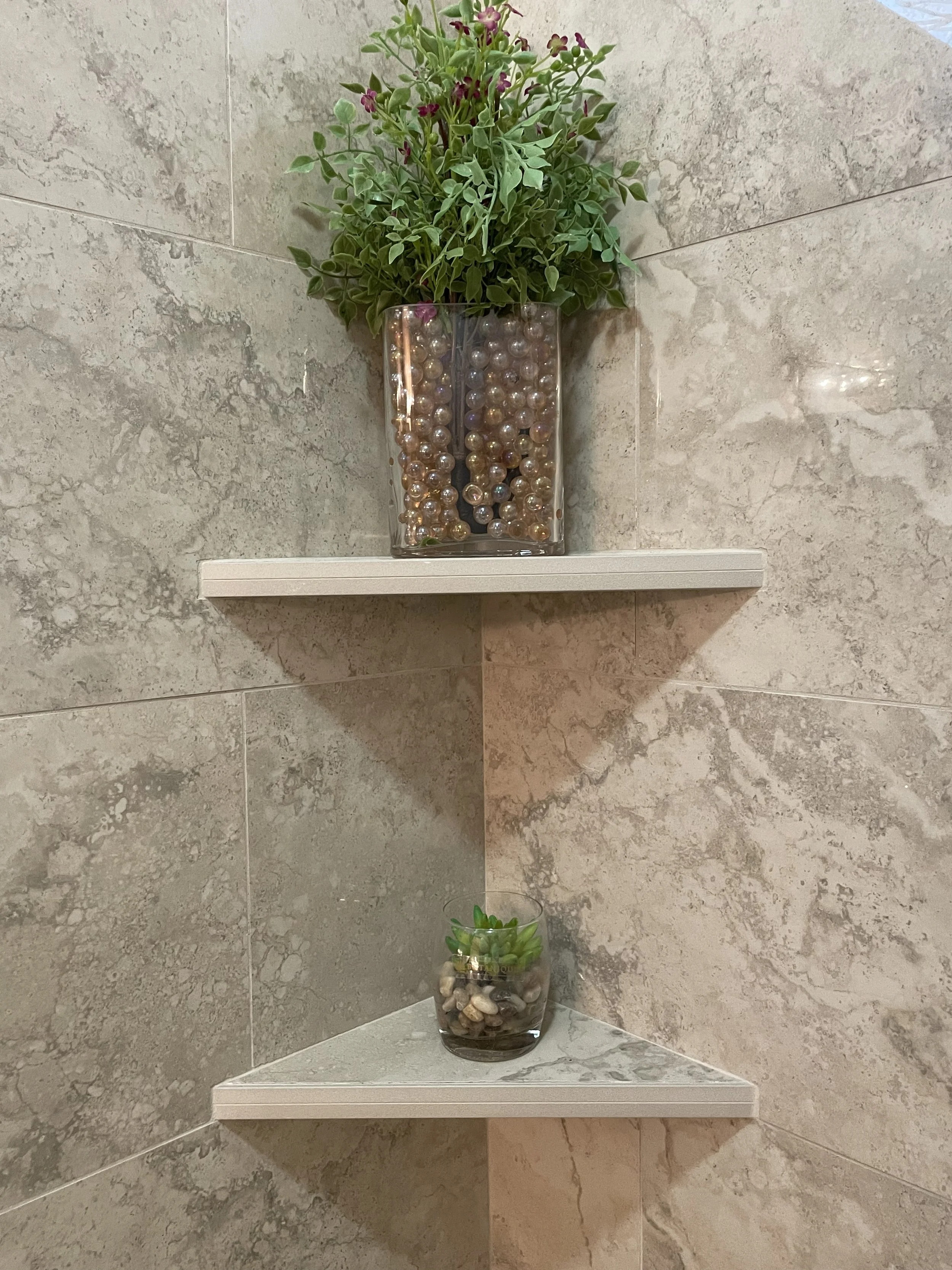 A decorative setup with two glass vases, one containing pearl-like beads and green leafy plants with small purple flowers, placed on a small marble shelf against a beige marble tiled wall.