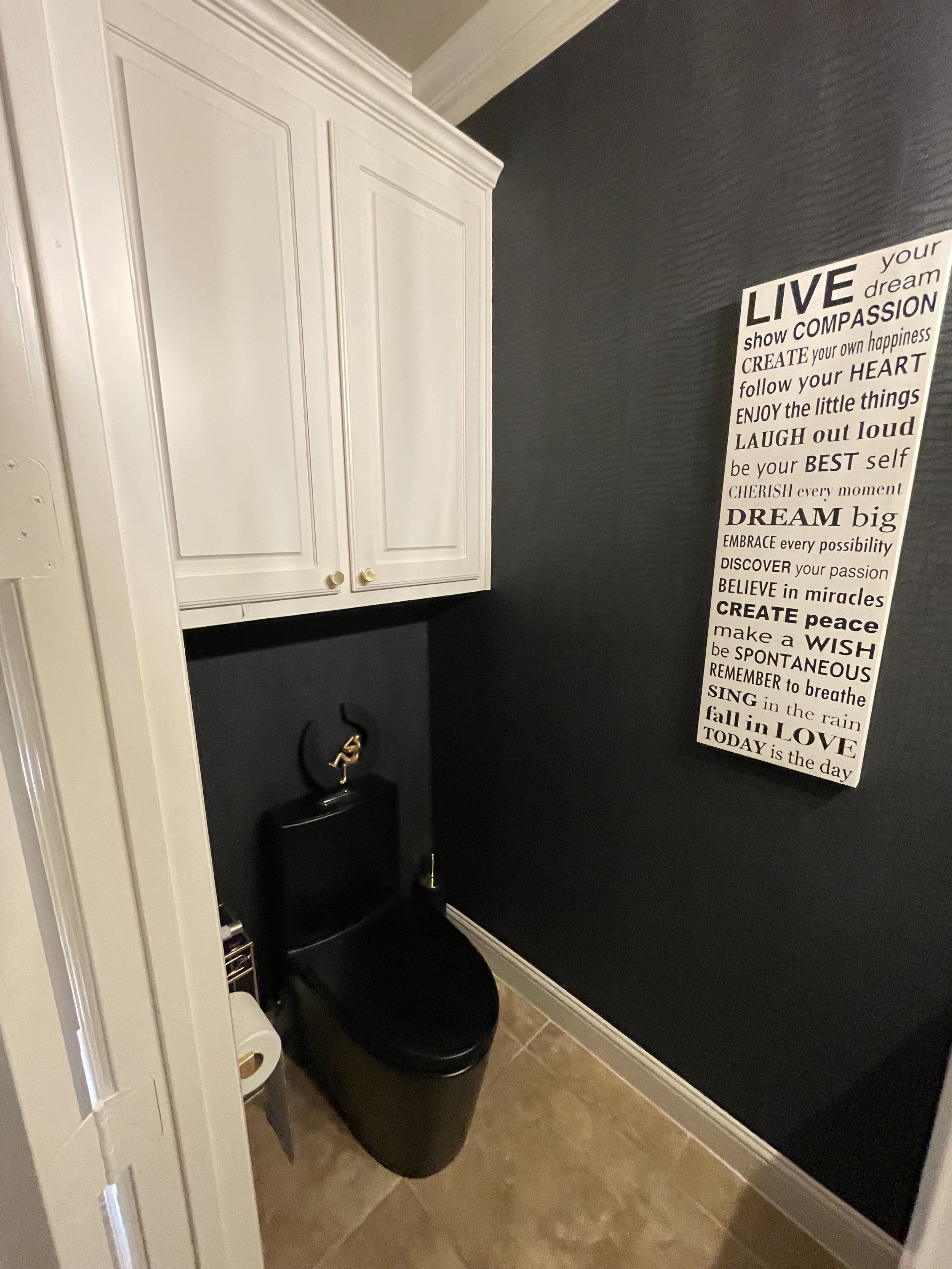 Bathroom corner with white cabinet, black toilet, a black tote bag, a wall with black textured paint, a white decorative frame with inspirational quote, and tiled flooring.
