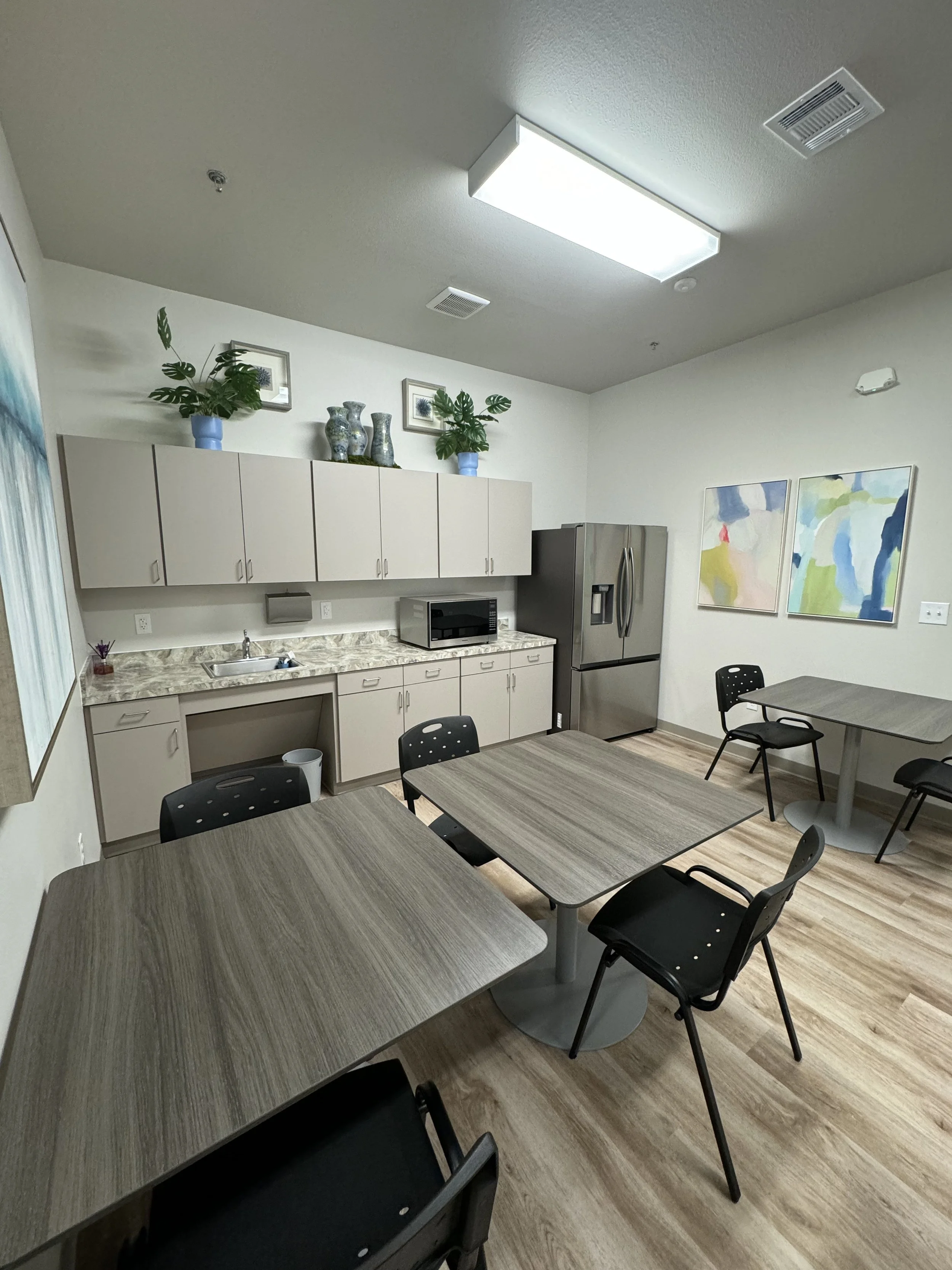 Office break room with tables and chairs, kitchen area with cabinets, a stainless steel refrigerator, a microwave, framed artwork, decorative vases, and plants on top of the cabinets.