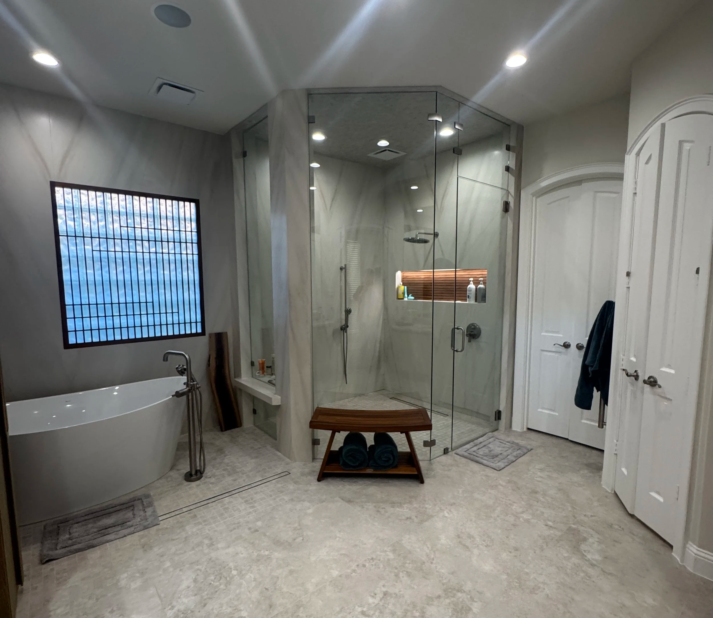 Modern bathroom with marble walls and floor, featuring a large mirror, glass shower stall, bathtub, window with grid pattern, and white doors.