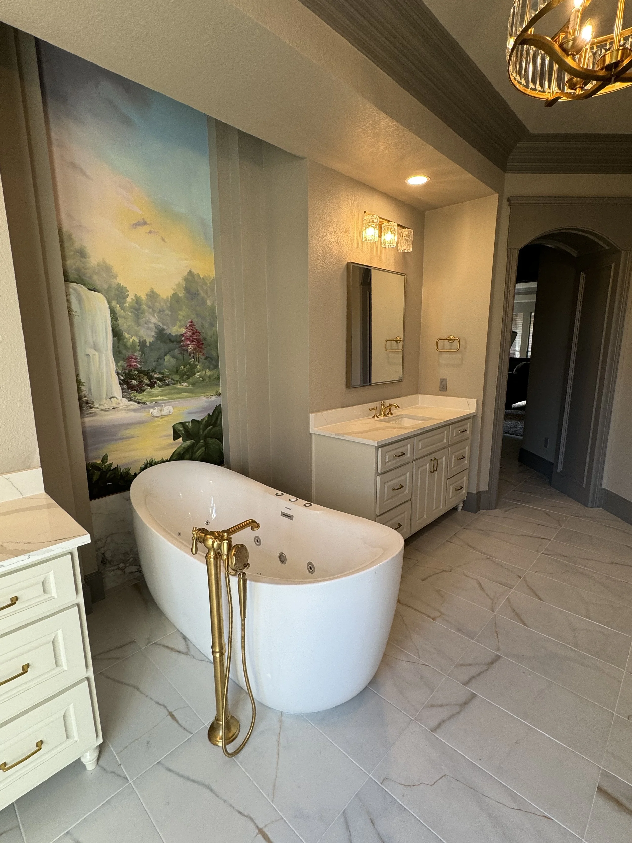 Bathroom with white freestanding bathtub, gold fixtures, a vanity with a mirror, a mural of a waterfall and landscape, and a chandelier.