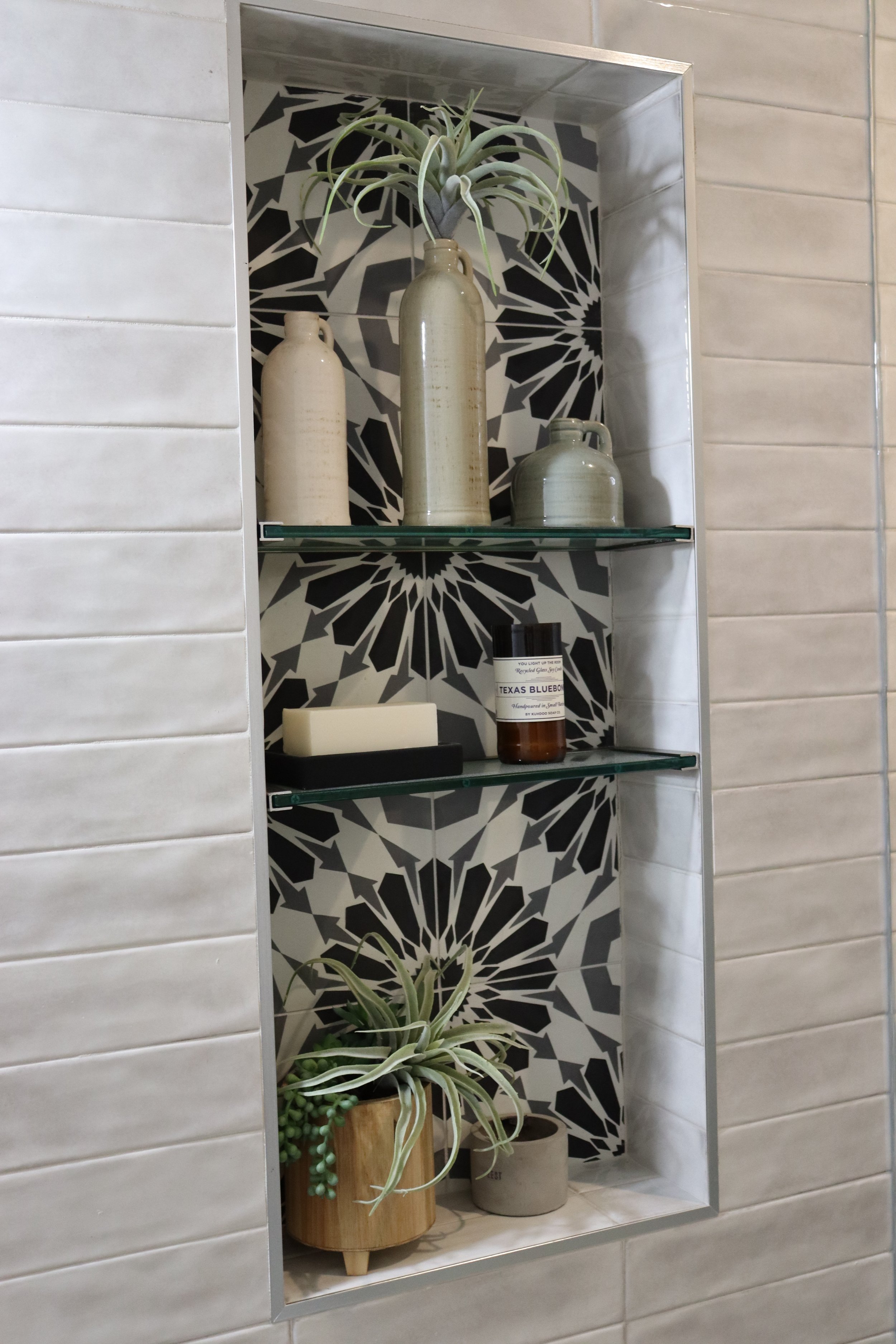 A built-in wall shelf with a black and white geometric tile background, holding three vases and a small plant. The top vase has a tall, narrow shape with a smaller, rounded vase beside it. The bottom shelf has a rectangular soap bar and a bottle of Texas Bluebonnet soap, with a potted plant featuring long, slender leaves trailing over the planter.