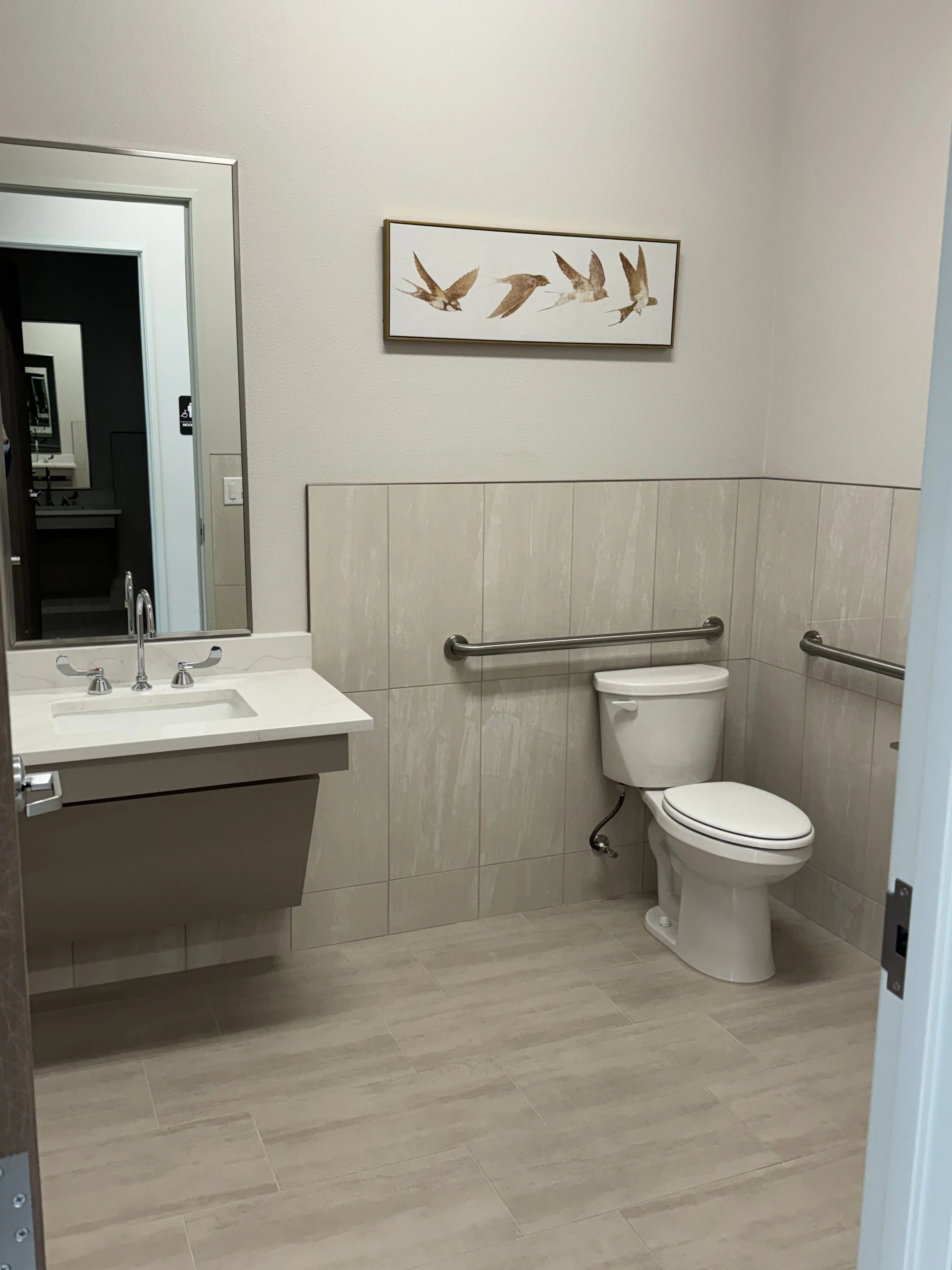 A clean restroom with beige tiled floor and half-tiled walls. A white sink with a silver faucet is on the left, reflected in a mirror. To the right, there is a white toilet with grab bars on the side walls. Above the toilet, a framed artwork of four flying birds is hung on the wall.