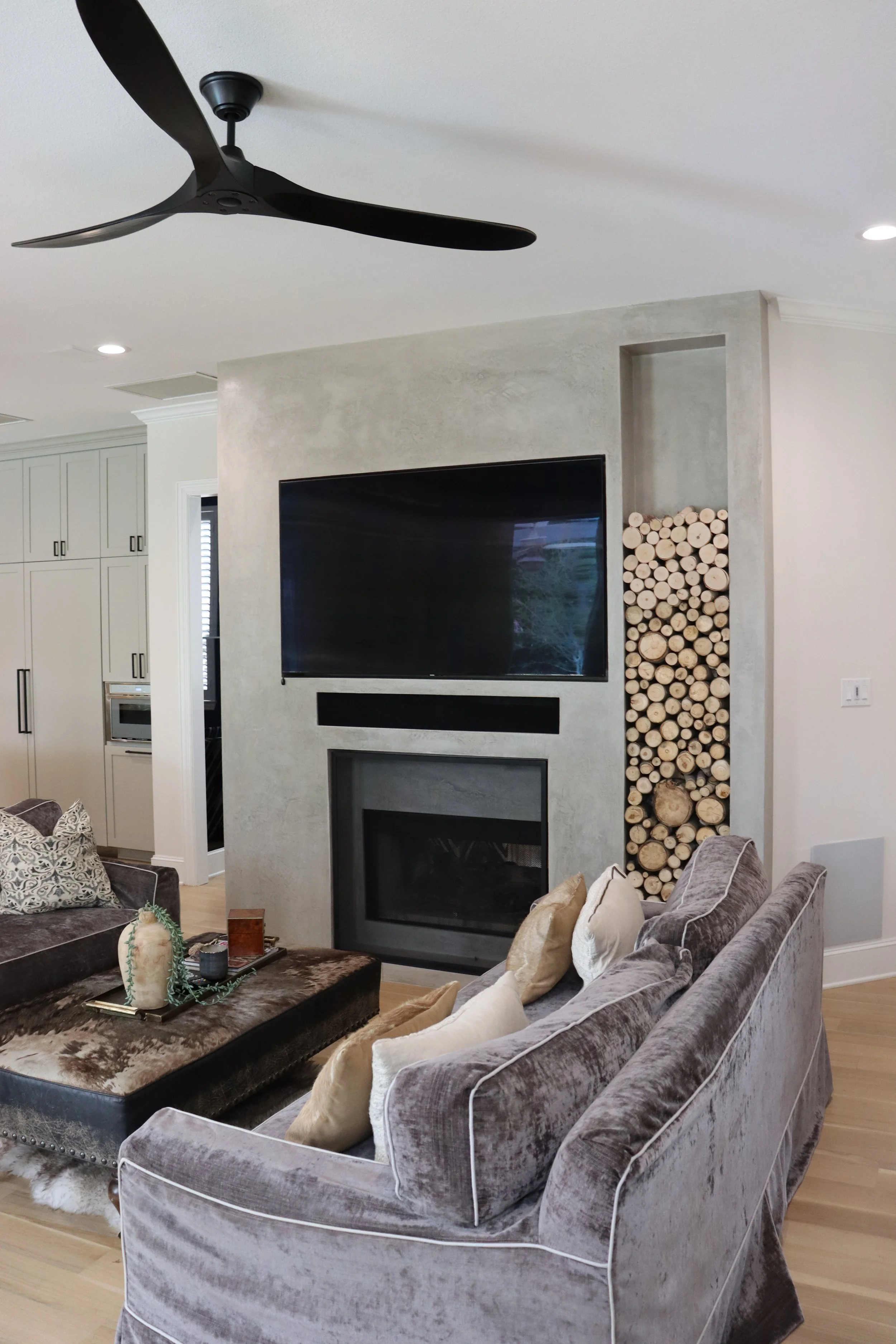 Living room with a gray velvet sofa, a black entertainment wall with a TV, and a stack of firewood in a niche. There is a ceiling fan and recessed lighting.