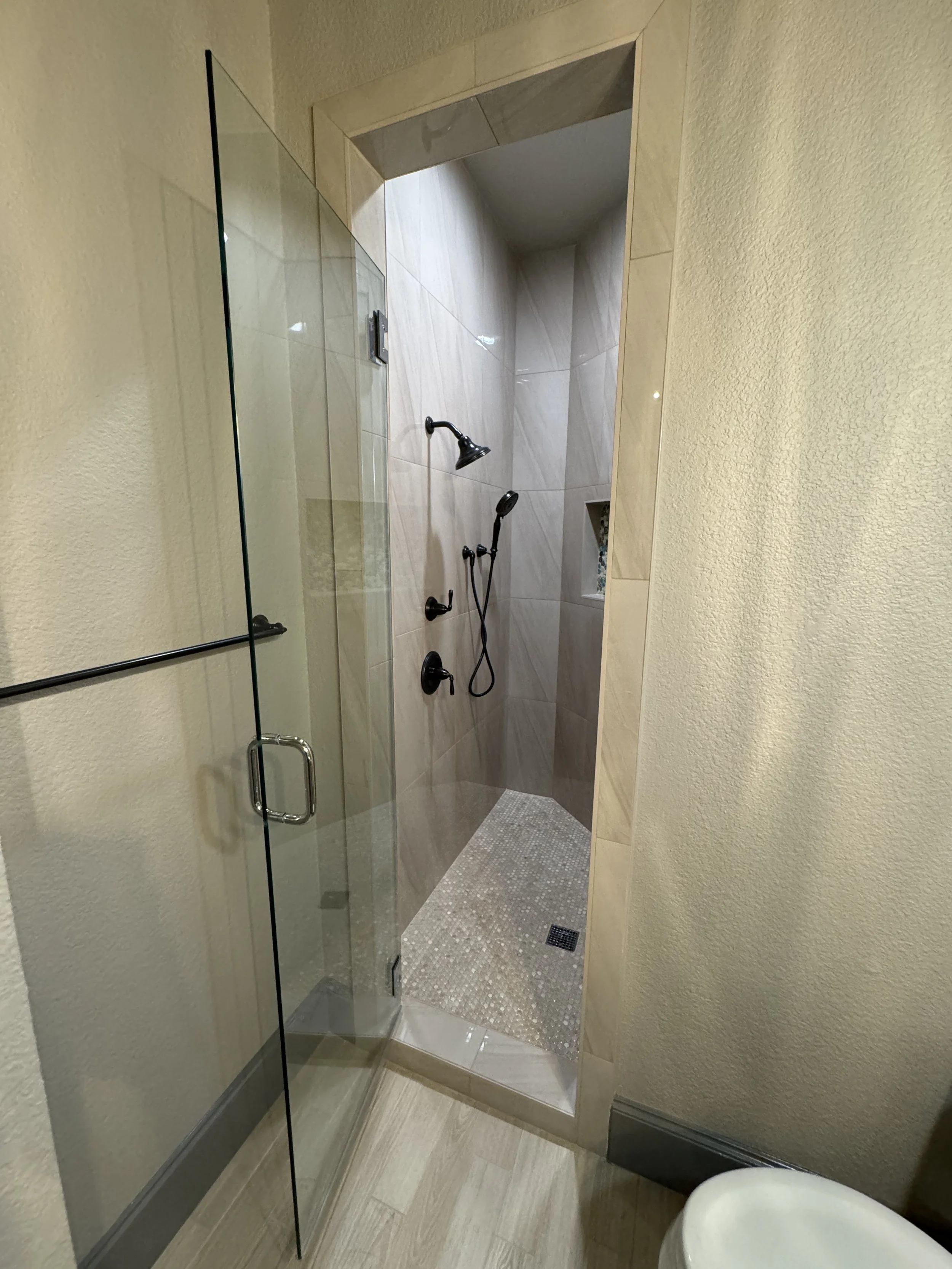 Small bathroom with glass shower enclosure, beige walls, and a corner shower with corner shelves, a showerhead, and a handheld shower.