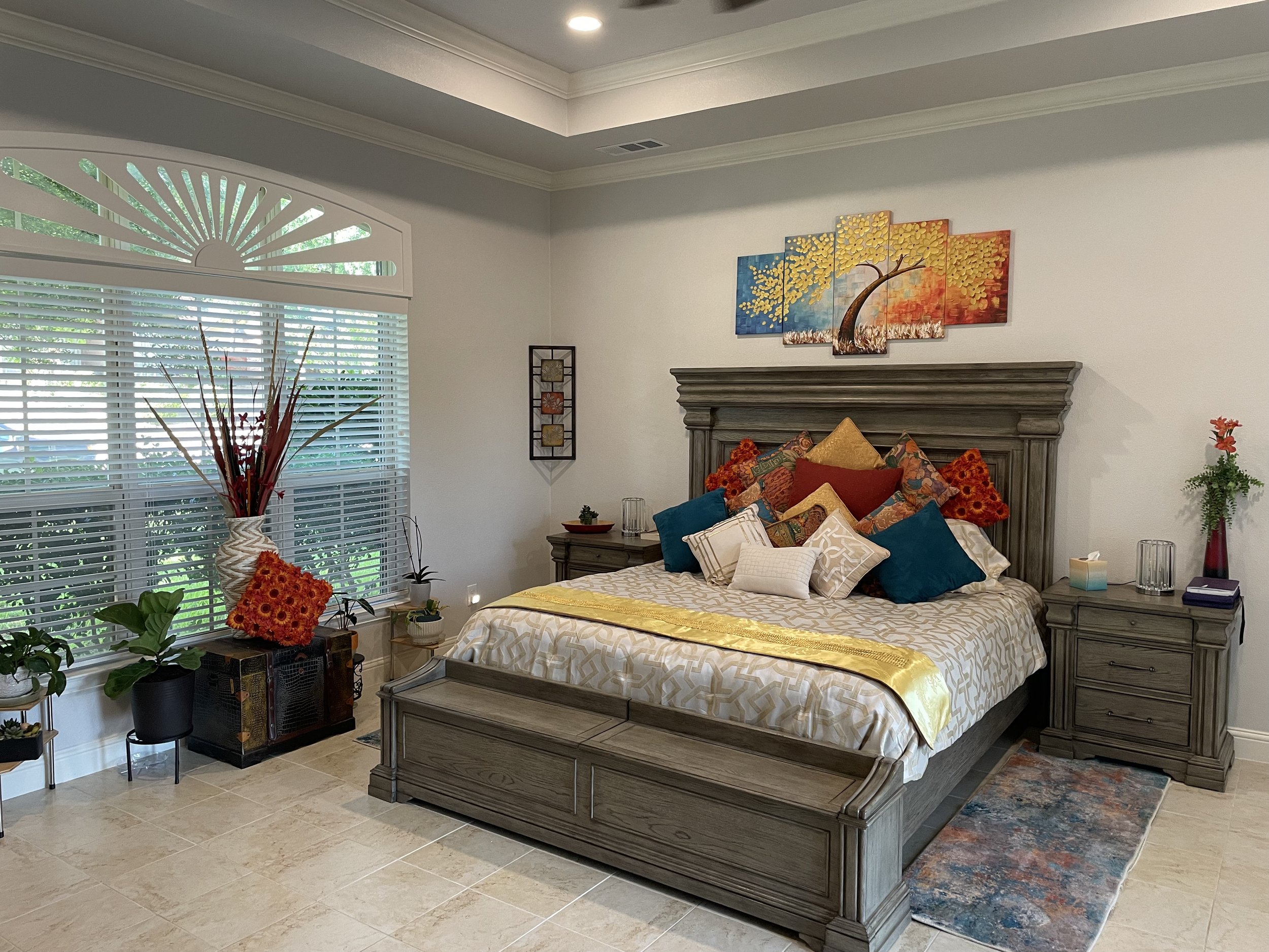 A bedroom with a large bed decorated with colorful pillows, two nightstands, and a mural of a tree on the wall above the bed. There are various plants and decorative items near the window and on the floor.