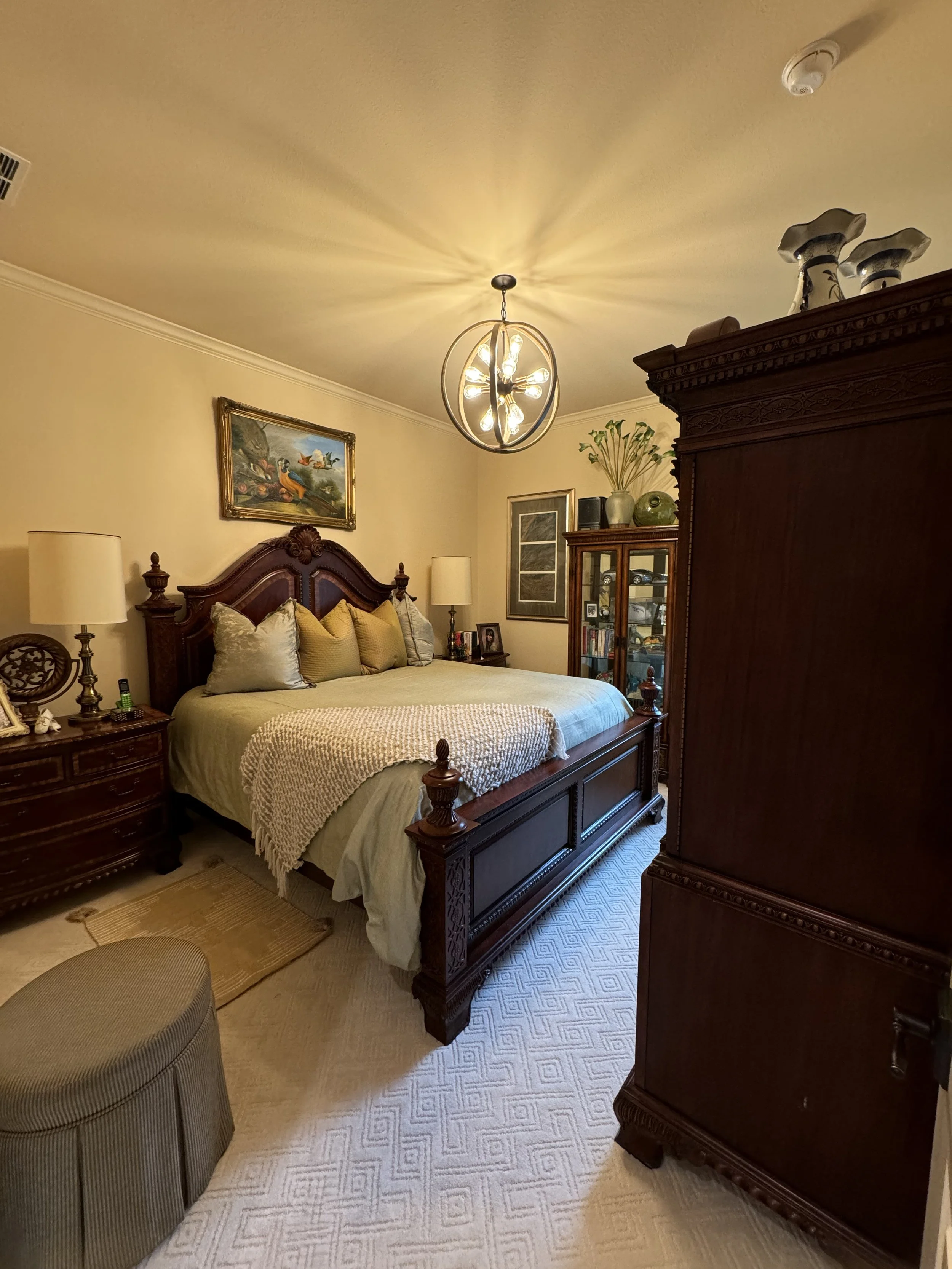 A bedroom with a large wooden bed, beige bedding and multiple pillows, side tables with lamps, a painting above the bed, a glass-fronted cabinet with decor items, and a dark wooden armoire, illuminated by a modern spherical chandelier, with a patterned carpet and beige walls.