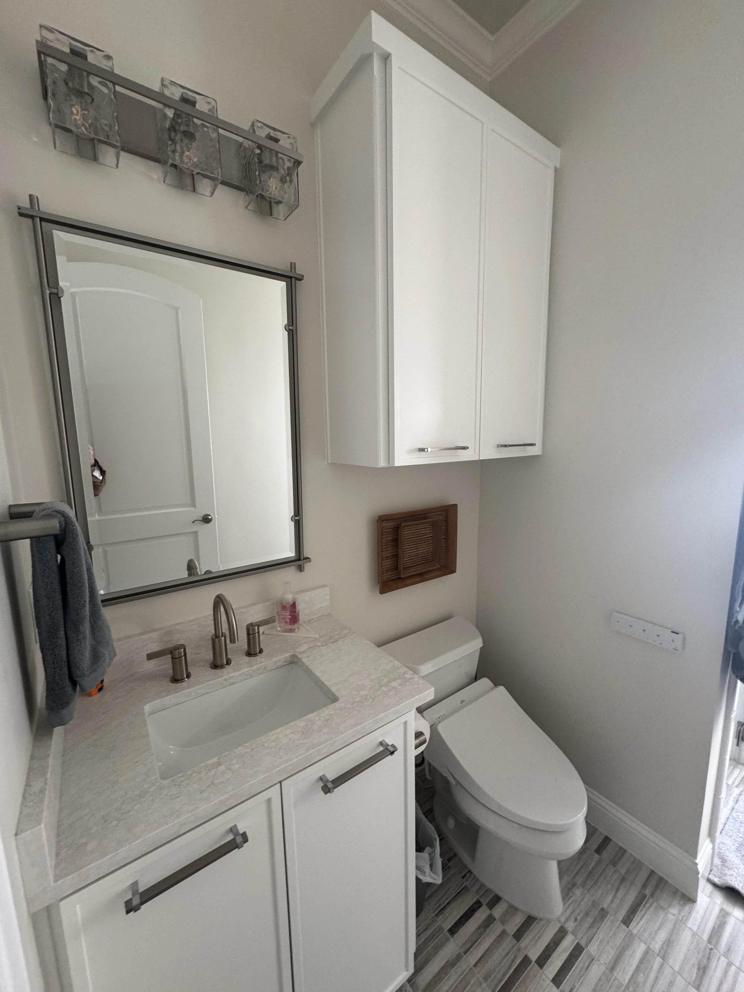 A small bathroom with white cabinetry, a bathroom mirror above the sink, a beige countertop, a white toilet, and a patterned tile floor.