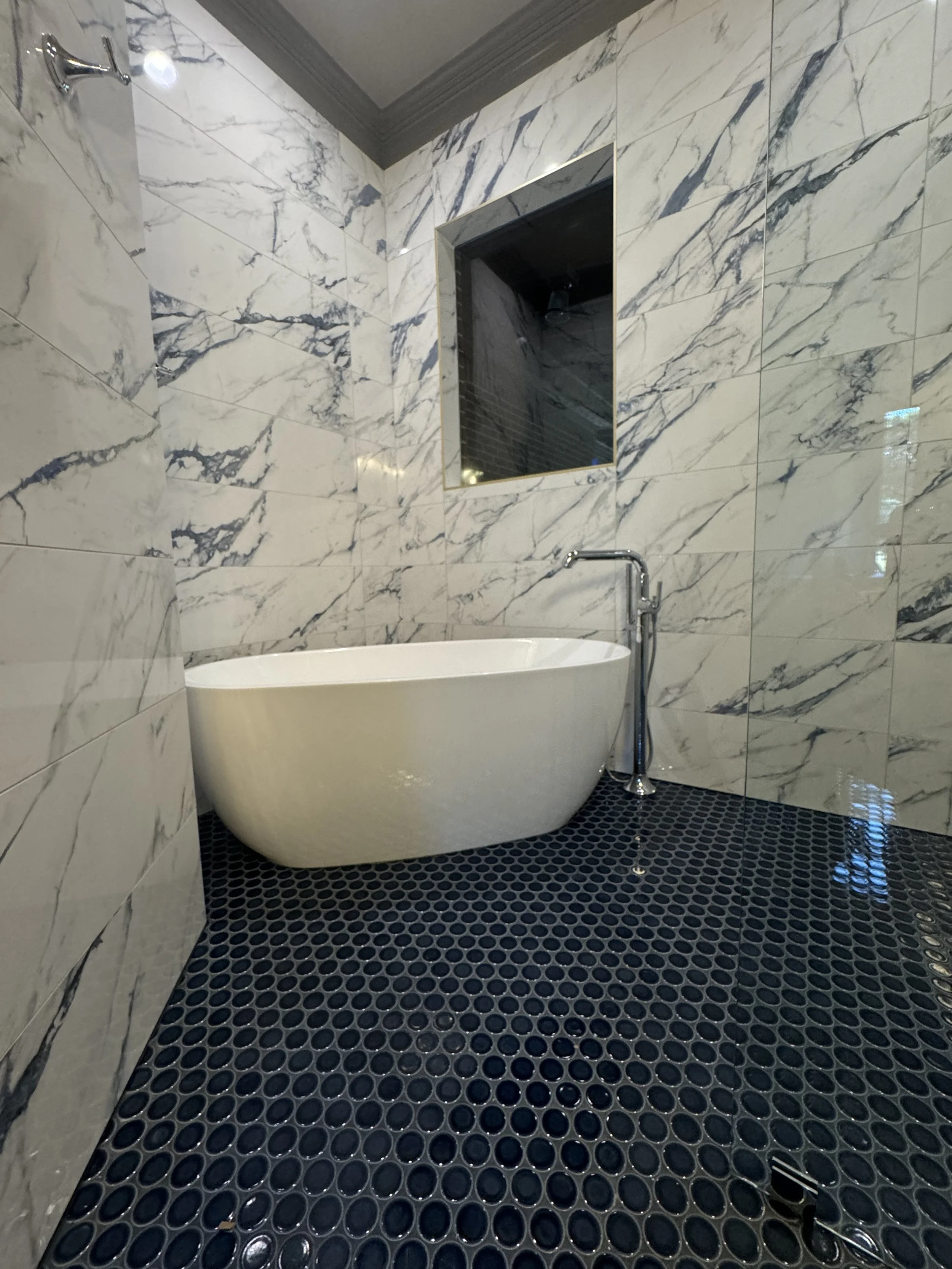 Modern bathroom with a bathtub, marble-tile walls, and a black hexagonal tile floor. There is a small rectangle mirror on the wall and a chrome faucet attached to the bathtub.