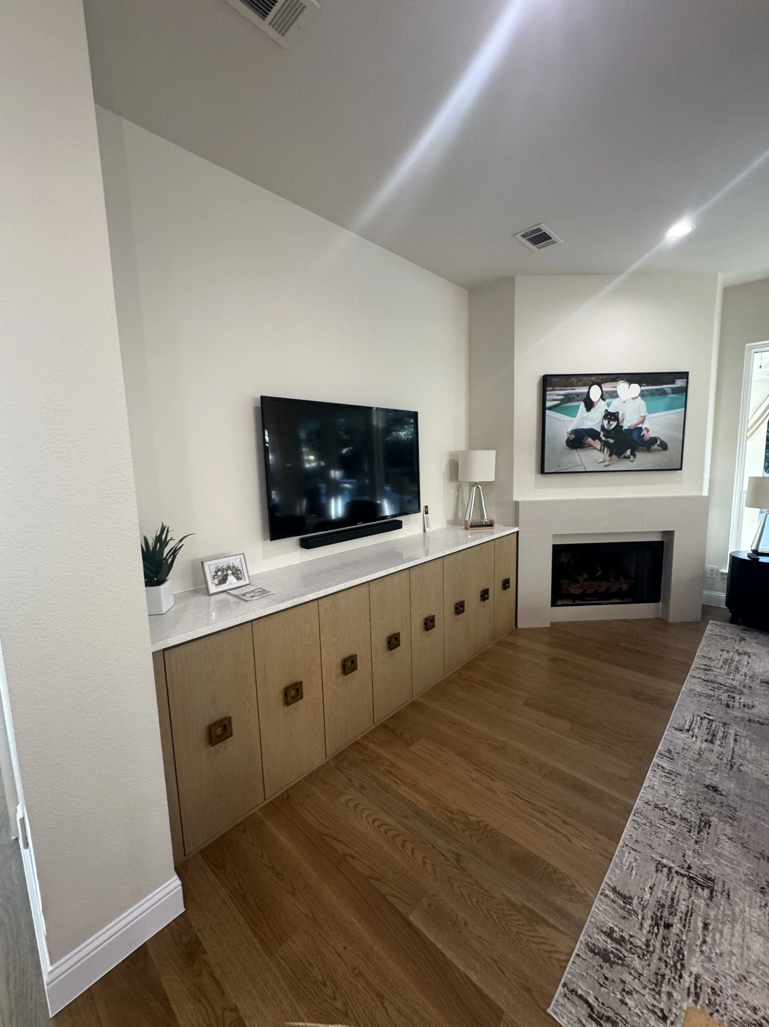 A living room with a wall-mounted flat-screen TV, a fireplace, a white cabinet with wooden doors, a small potted plant, a framed photo, a table lamp, a rug, and a window with sunlight.