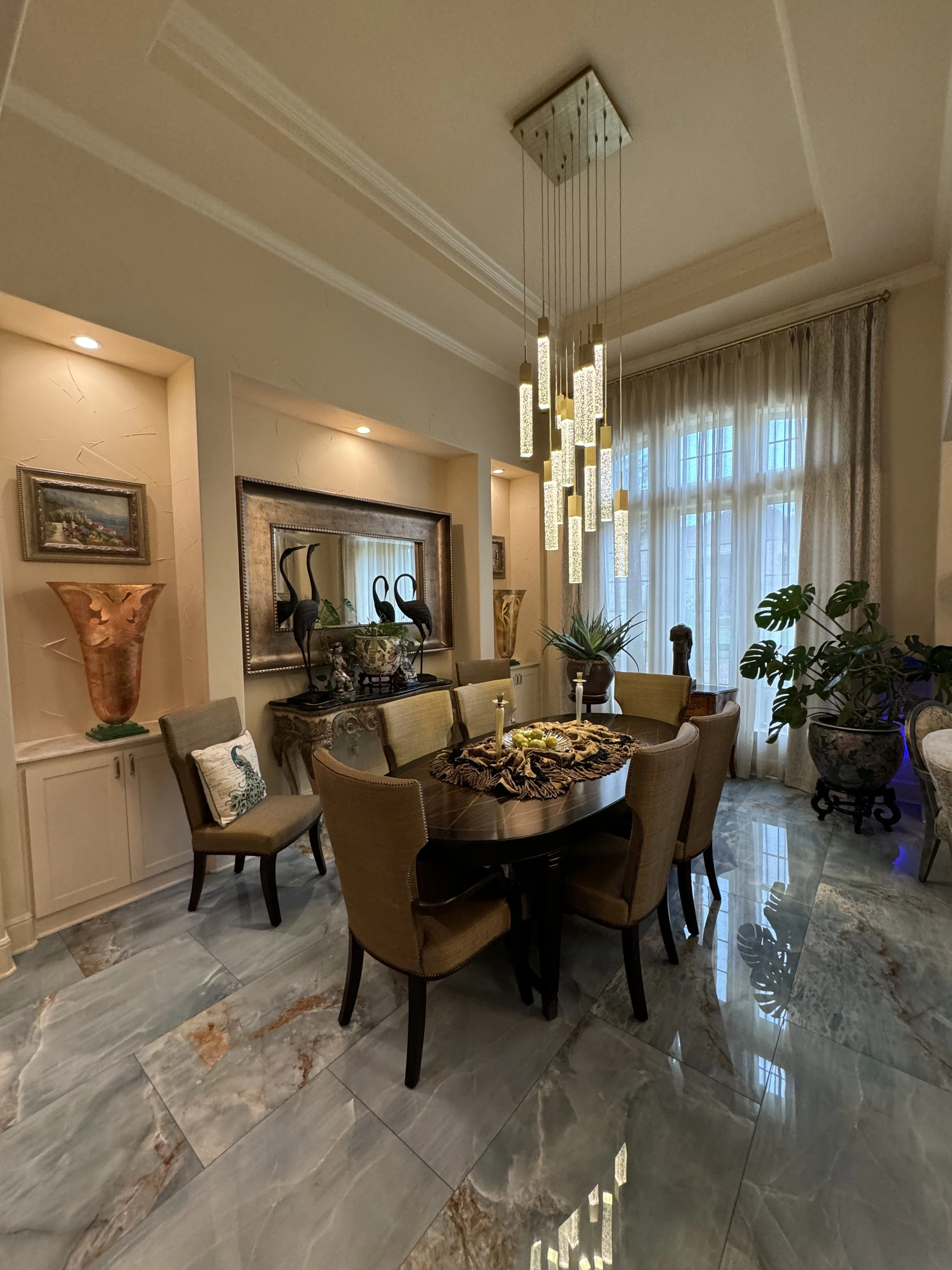 Photo of a dining room with a round wooden table, upholstered chairs, large window with sheer curtains, decorative vases, and a modern hanging chandelier.
