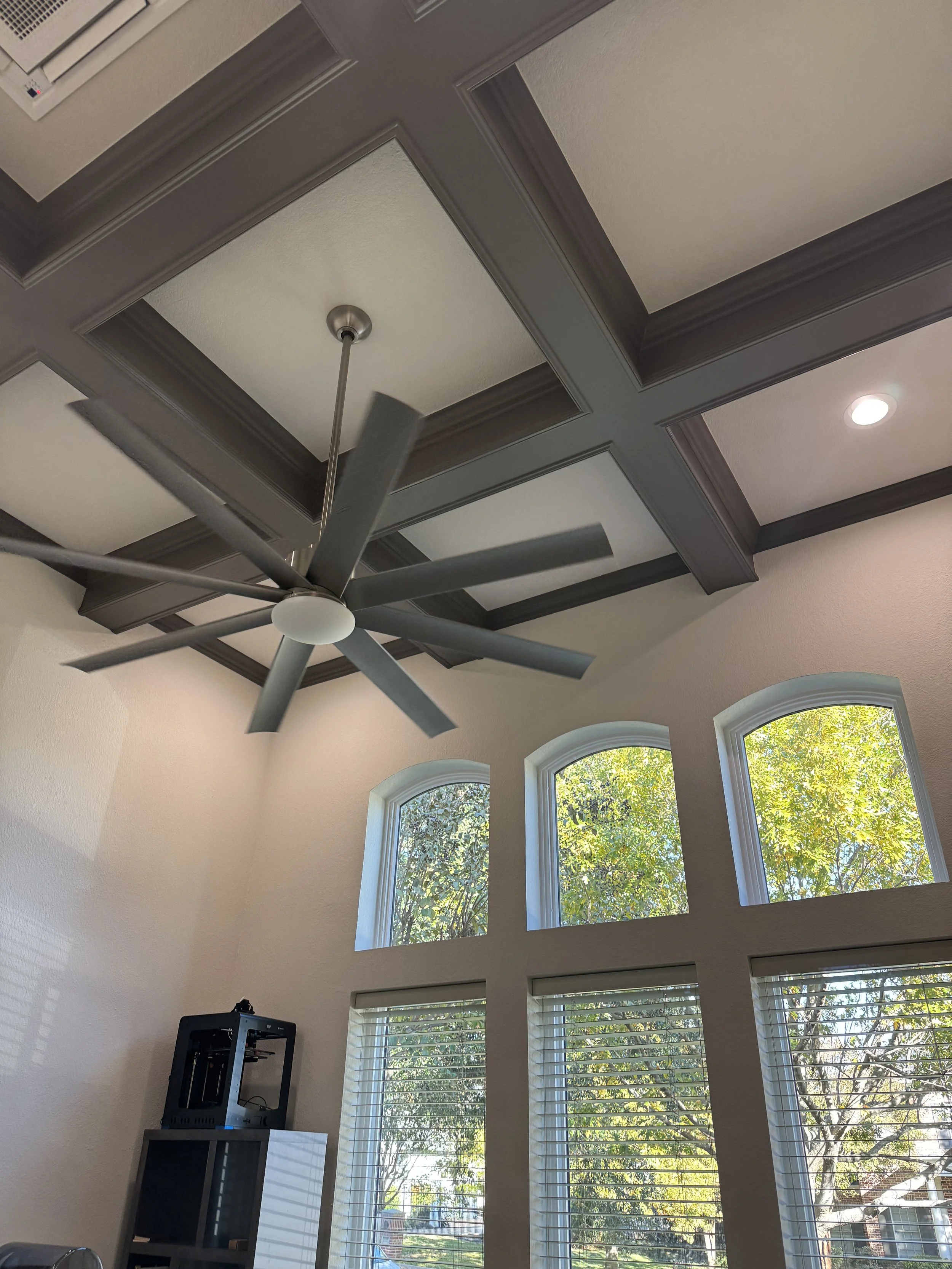 A room with high ceiling featuring a ceiling fan with multiple blades and a light, large windows with blinds showing trees and outdoor scenery, and a black 3D printer on a table.