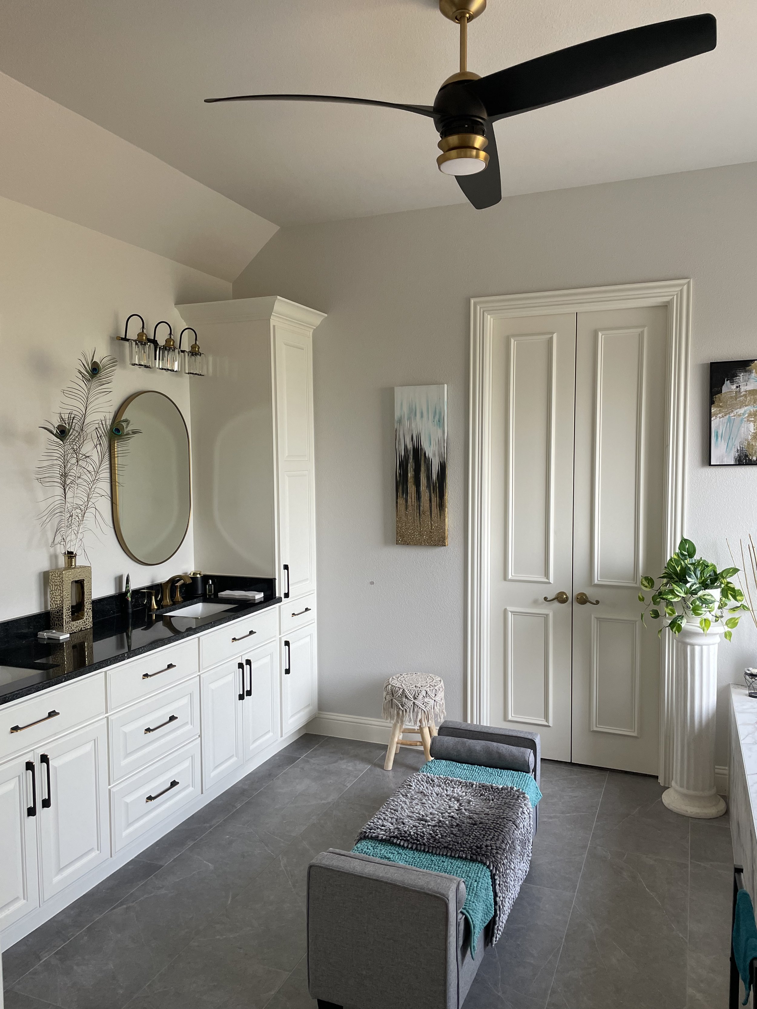 A modern interior space with a white cabinetry vanity, a wall mirror, decorative items, and artwork. There is a ceiling fan with black blades and gold accents, and a potted plant in a white pedestal. The room has a gray tiled floor and a door with detailed molding.