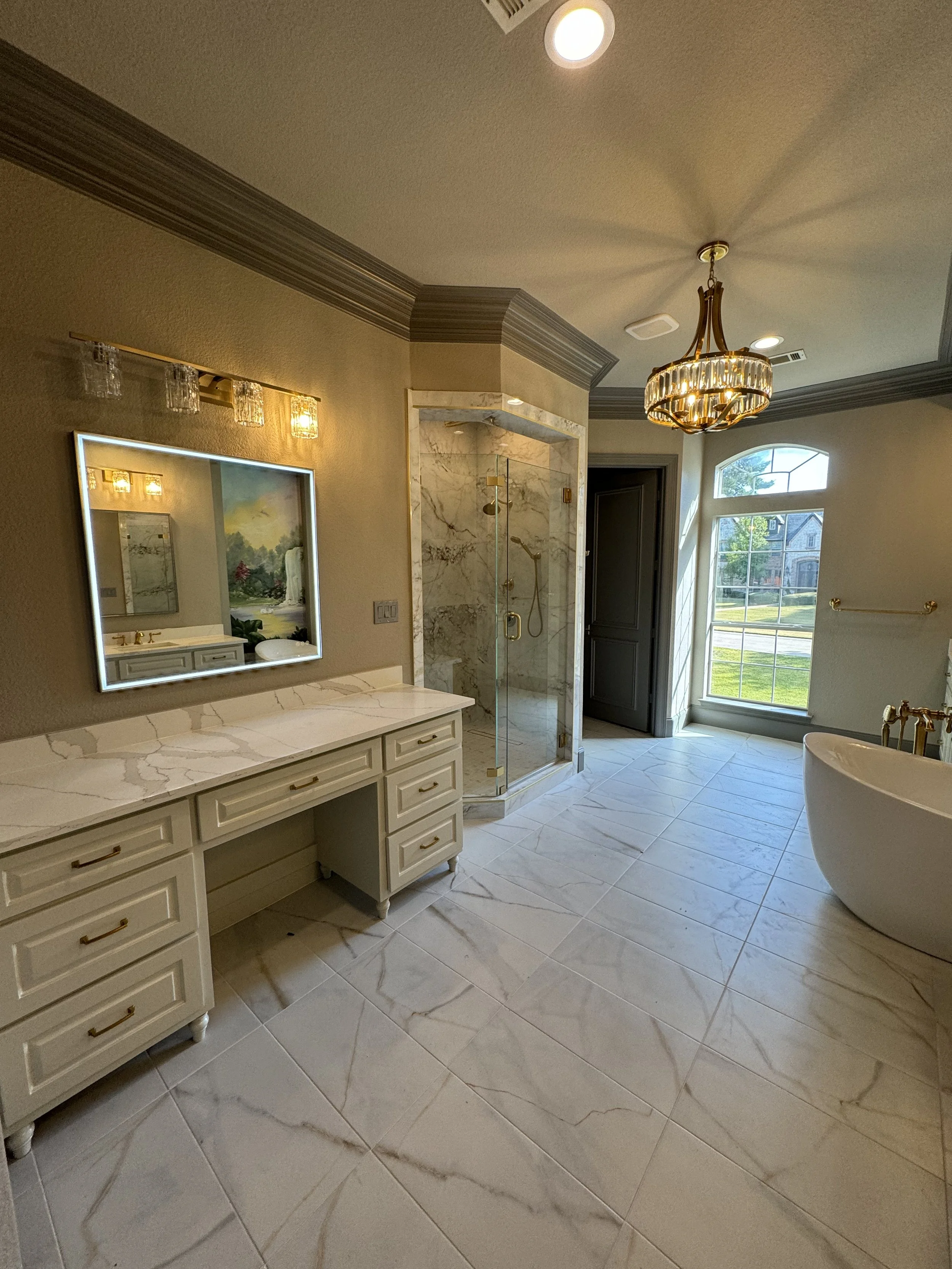 Bathroom with tiled floor, vanity with marble countertop and mirror, glass-enclosed shower with marble walls, freestanding bathtub, window with sunlight, chandelier light fixture, and small wall-mounted light fixture.