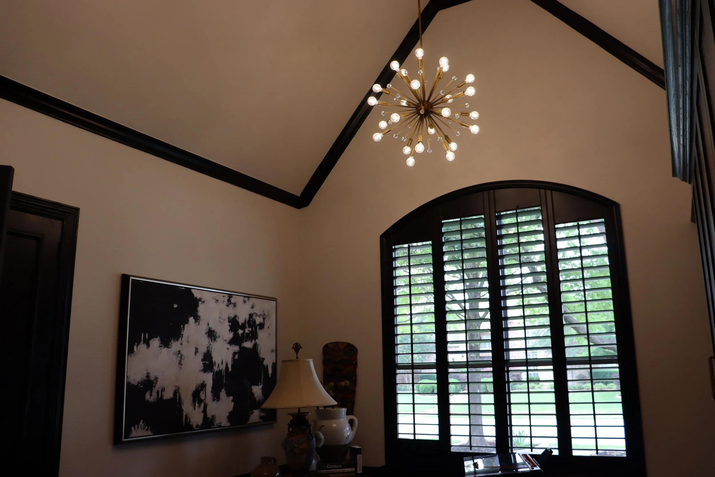 A room with a decorative chandelier hanging from the ceiling, window with shutters, and various art pieces and vases on a table.