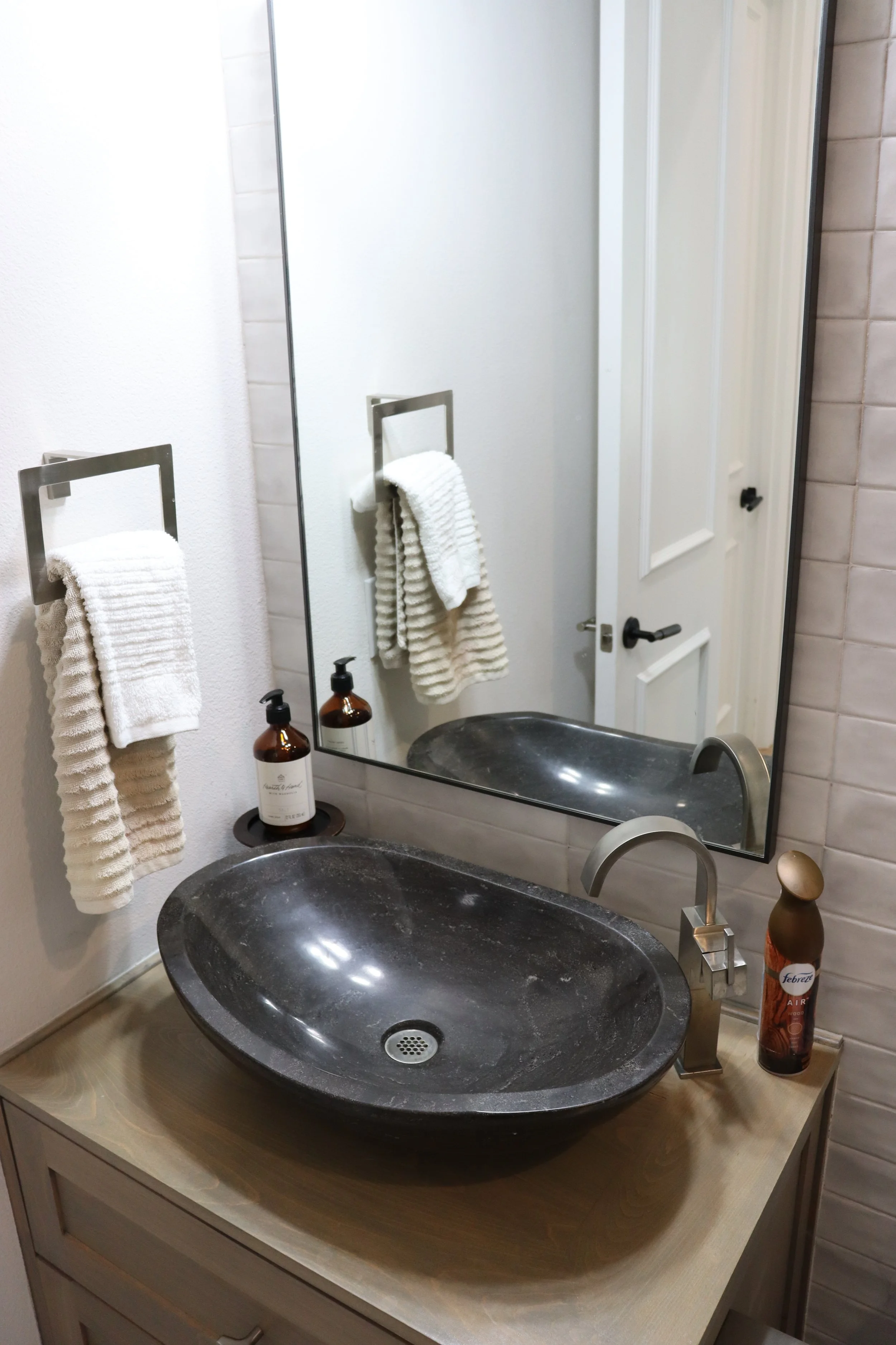 Bathroom sink with black countertop, oval sink, gold faucet, two bottles of soap, a mirror, a white towel on a silver towel ring, closed white cabinet door, and a soap bottle on the light tan wooden vanity.