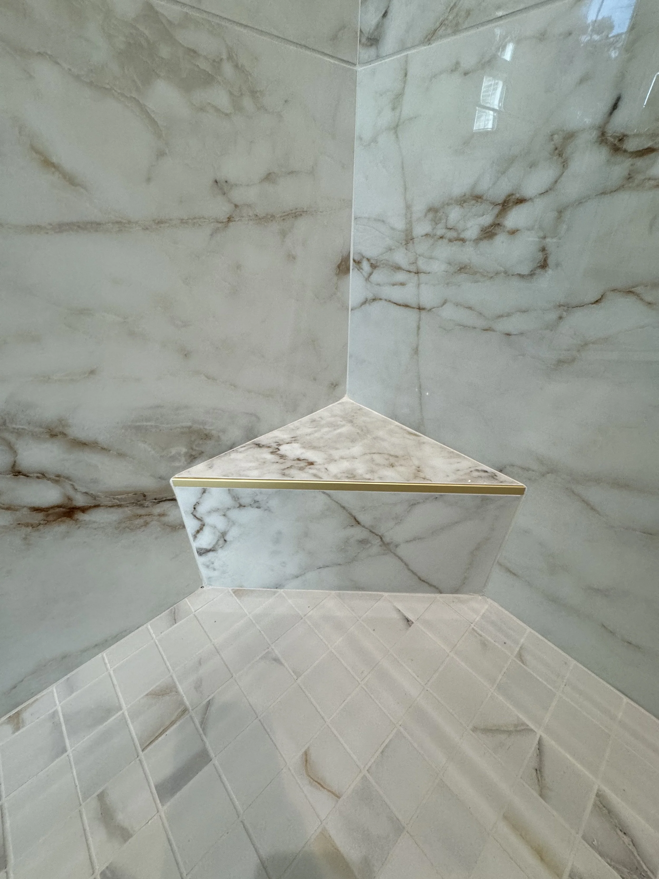 Corner of a bathroom with marble walls and tiled floor, featuring a triangular marble shelf with a gold trim.