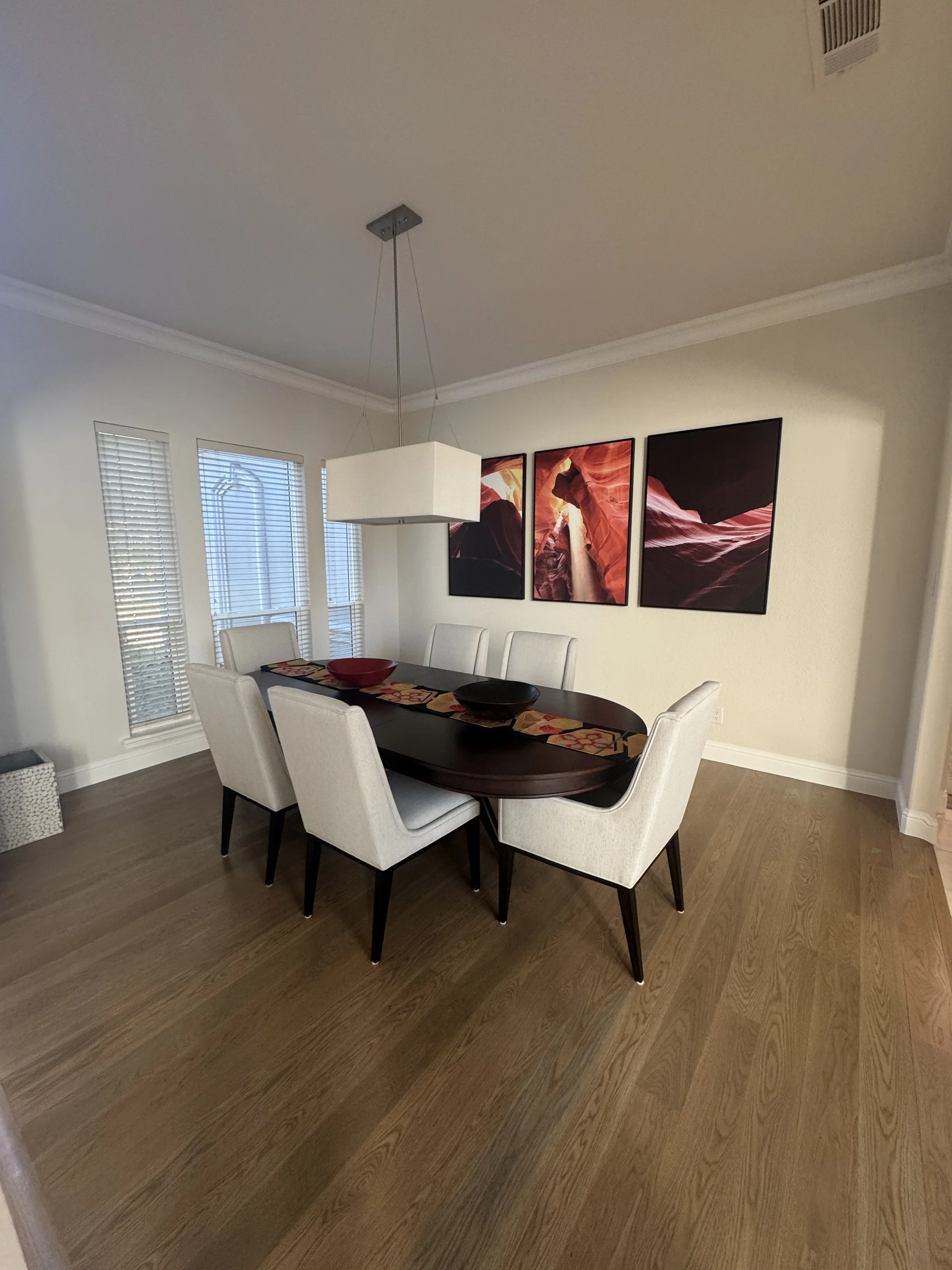 A dining room with a dark wood table, six cream-colored chairs, three window blinds, and three framed landscape pictures on the wall. There is a hanging white rectangular light fixture over the table.