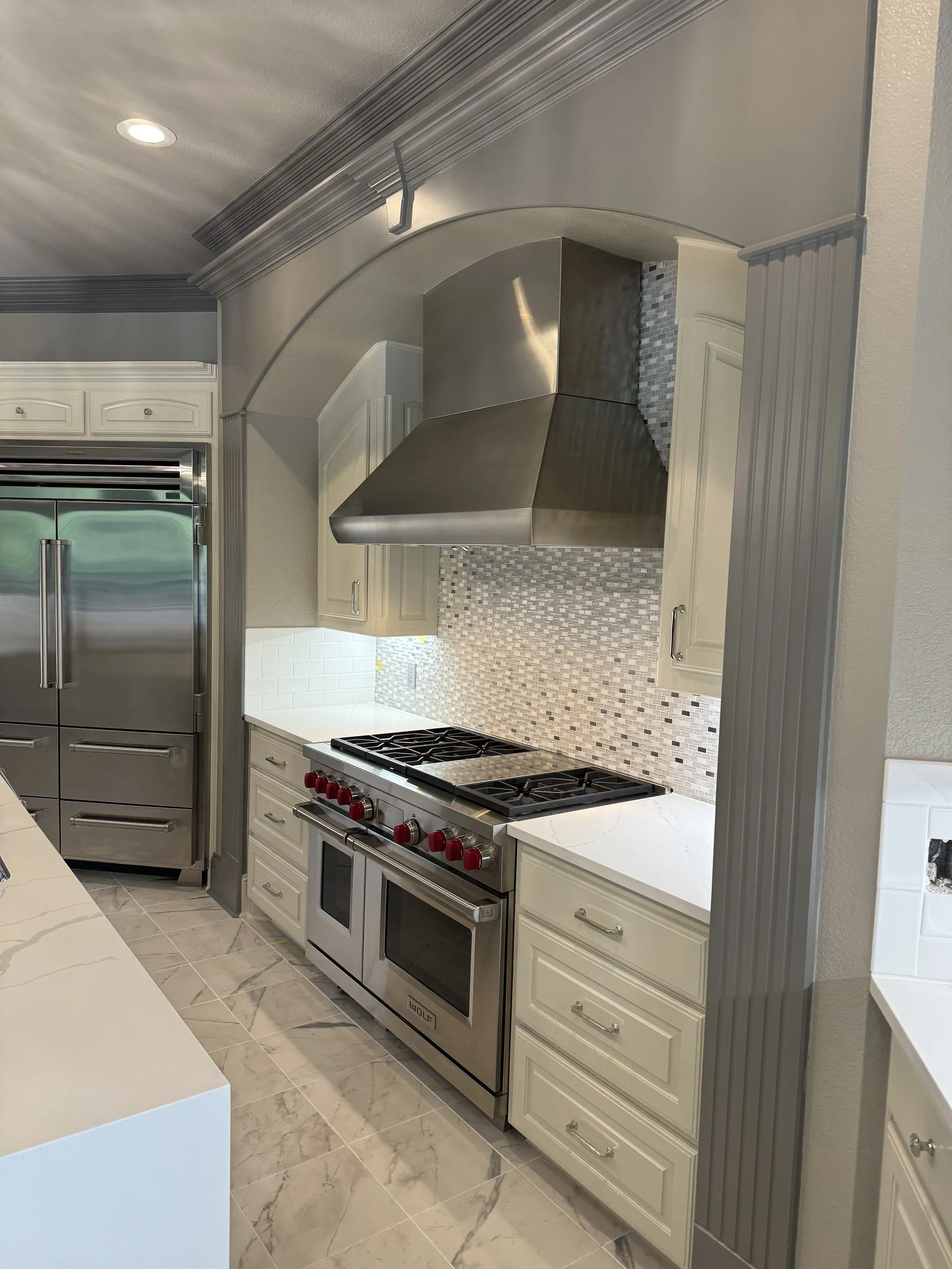 Kitchen with stainless steel Wolf stove and oven, white cabinetry, mosaic tile backsplash, and gray decorative molding during daytime.