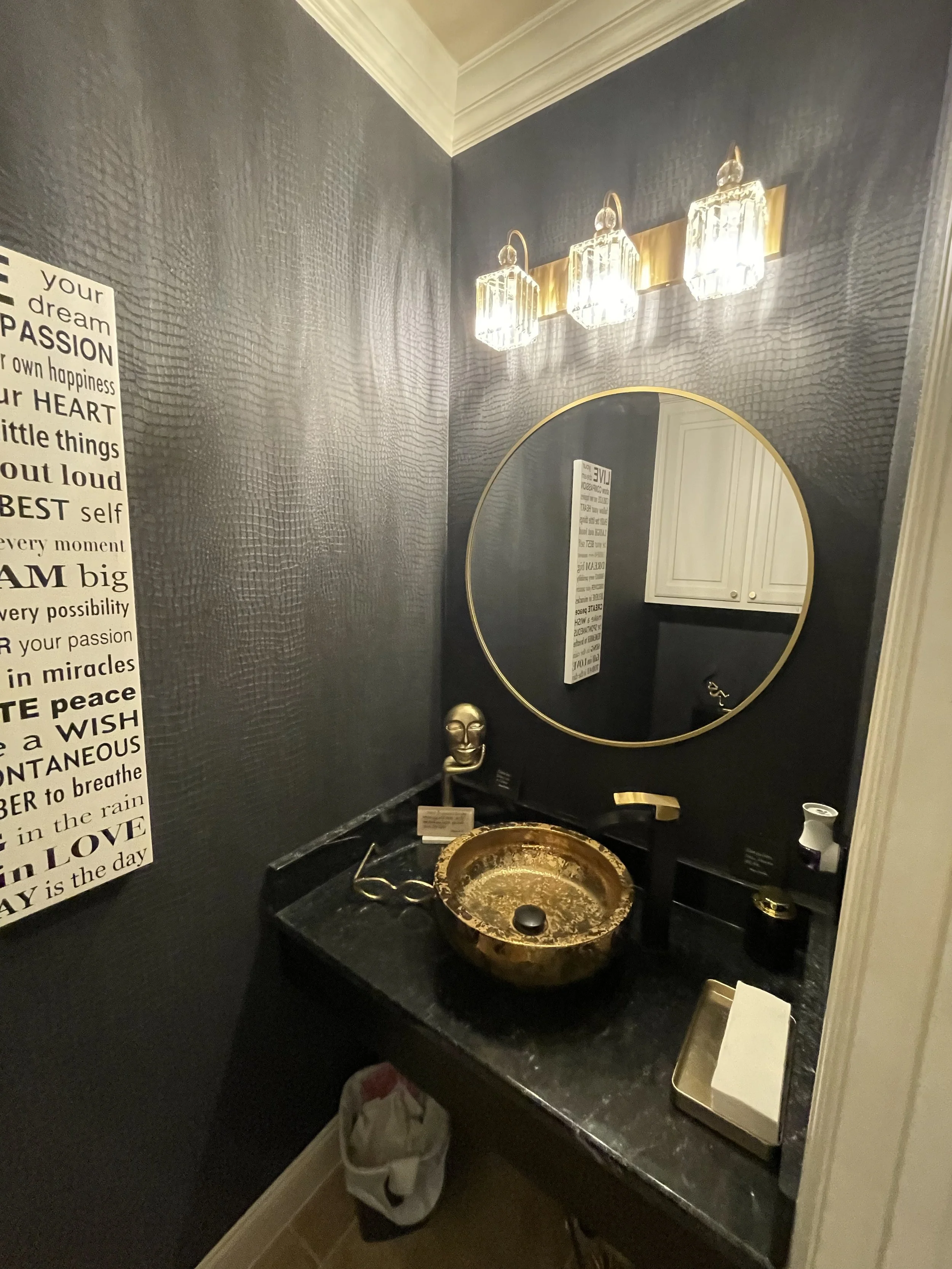 A bathroom vanity with a black countertop, gold and black fixtures, a round decorative bowl sink, a small gold bust sculpture, a rectangular mirror with a gold frame, and three crystal wall lights. There is a white cabinet reflected in the mirror, a wall with textured dark wallpaper, and a sign with inspirational words on the left side.