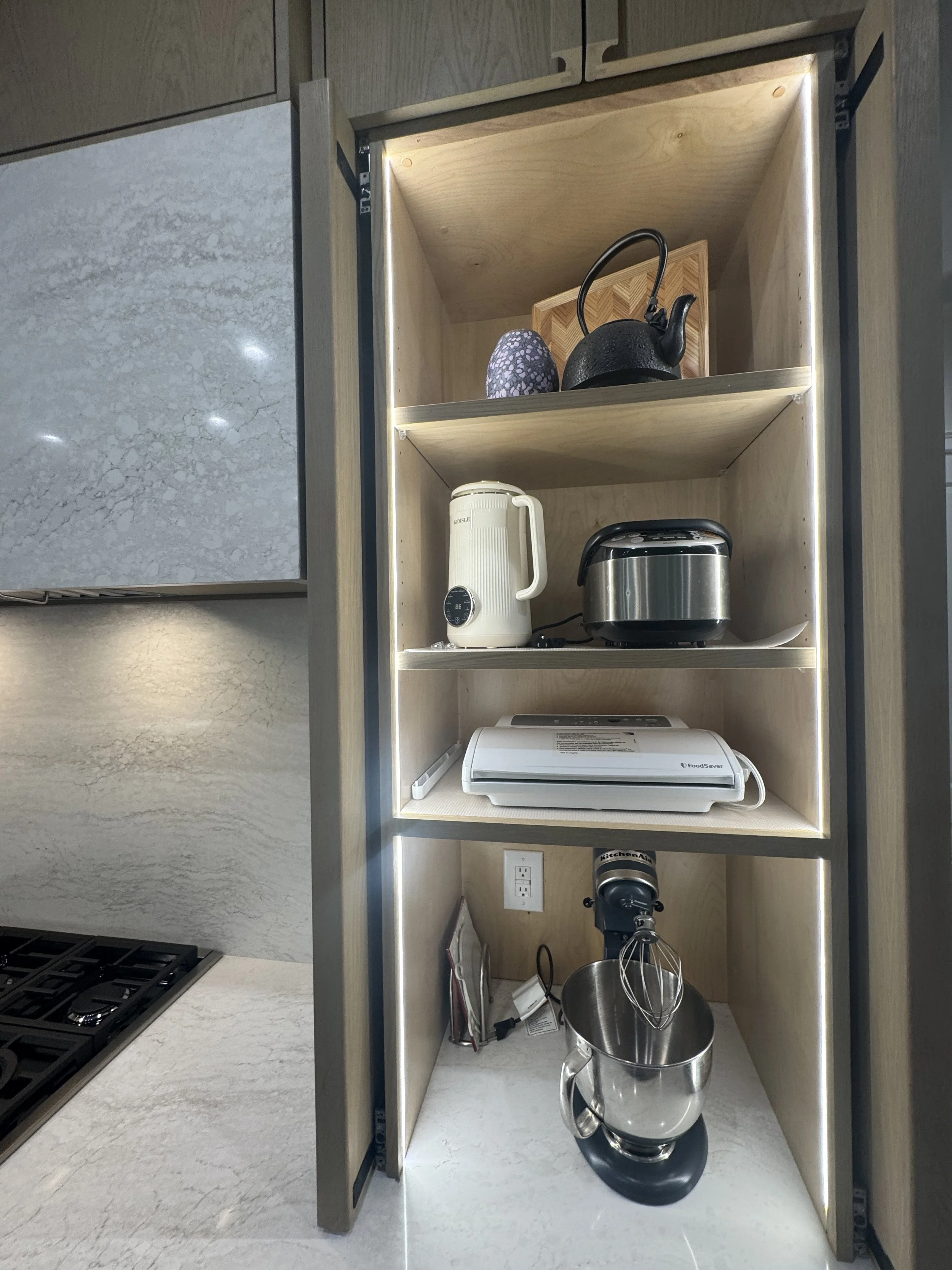 Open light wood kitchen cabinet with shelves holding a tea kettle, a decorative vase, a toaster, and a rice cooker, with a stovetop nearby.